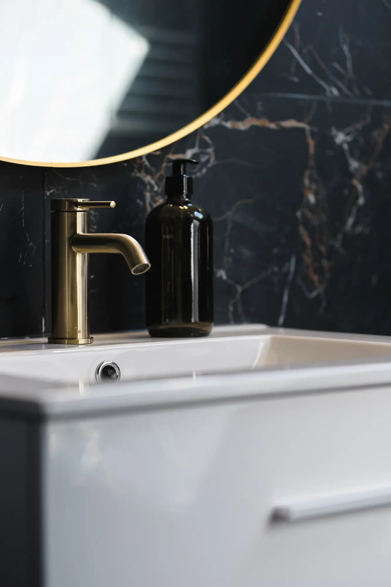 Vasque de salle de bain blanche avec robinet en laiton, bouteille noire de savon ou lotion, miroir rond au cadre doré, mur en marbre noir.