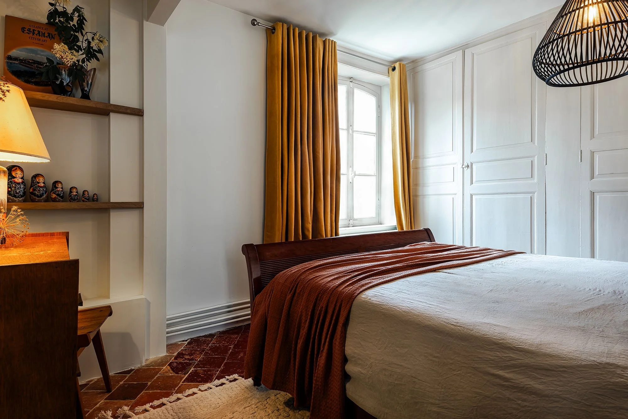 Chambre avec lit en bois, drap blanc et couverture marron, fenêtres avec rideaux marron, mur blanc, étagère avec décorations, lampe et tapis beige.