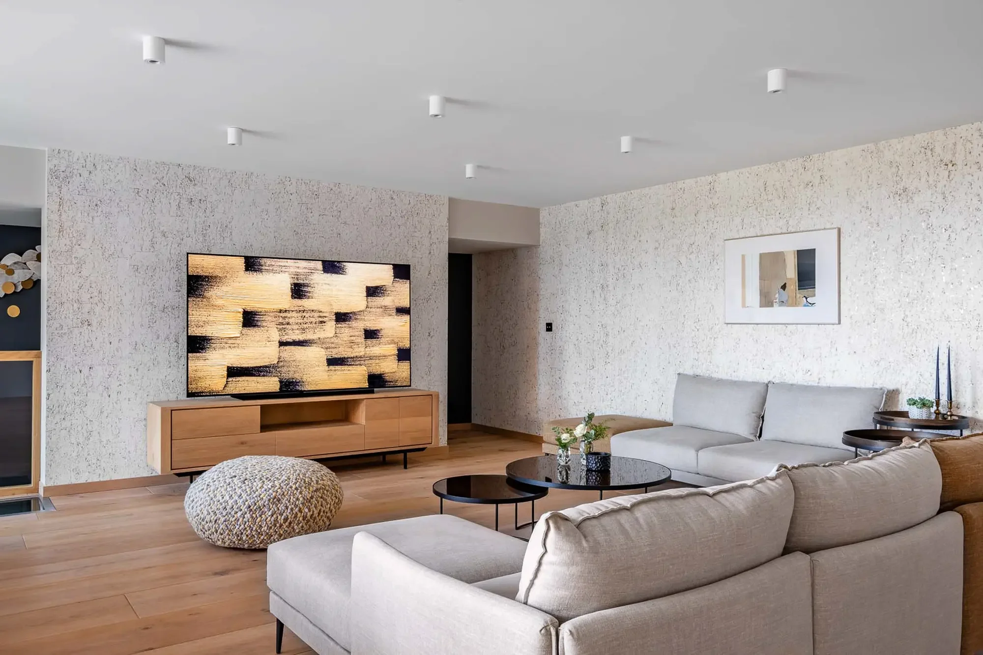 Salon moderne avec canapé beige, table basse ronde noire, pouf en laine textured, mur en pierre, télévision sur meuble en bois, lampe de plafond discrète, cadre photo sur le mur, plantes et bougies sur la table latérale