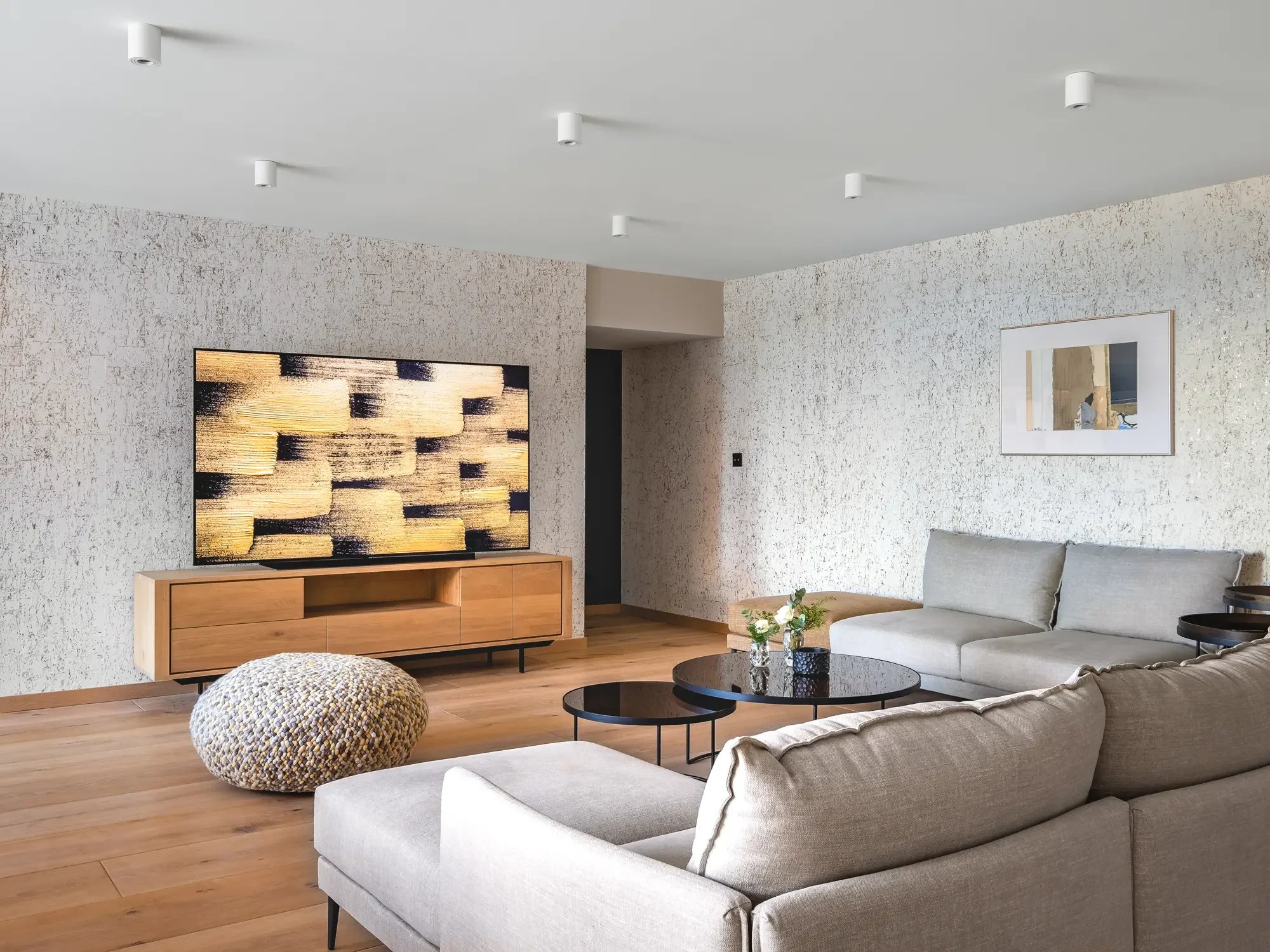 Salon moderne avec canapé beige, table basse ronde noire, mur en texture claire, télévision à meuble en bois, tableau abstrait, pouf en tissu multicolore, plantes et décoration.