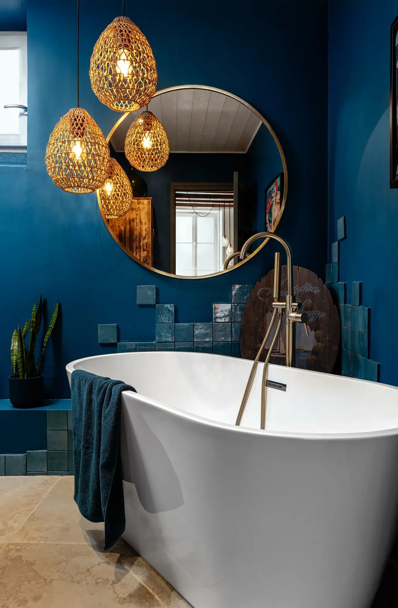 Baignoire blanche dans une salle de bain avec murs bleus, grands miroirs ronds, éclairage en suspension en rotin et plantes vertes.
