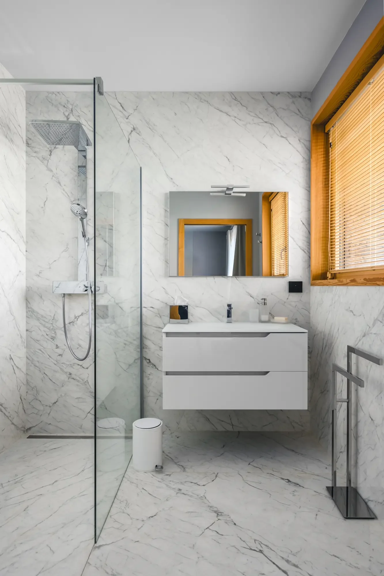 Salle de bain moderne avec murs et sol en marbre, lavabo blanc sous un miroir rectangulaire, fenêtre avec volets en bois, douche à l'italienne avec paroi en verre, accessoires en métal.