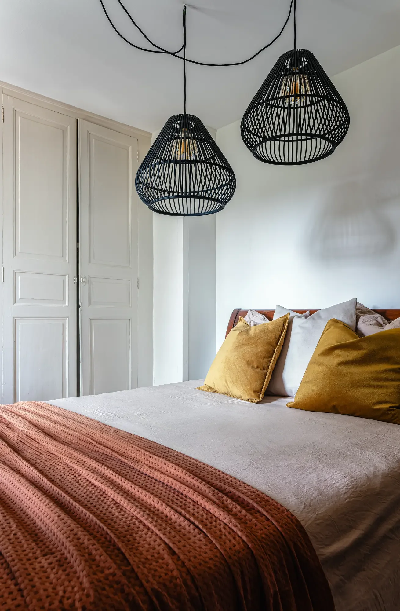 Chambre à coucher avec un lit avec draps beige, oreillers jaunes et blancs, et une couverture marron. Deux suspensions en rotin noir suspendues au plafond.