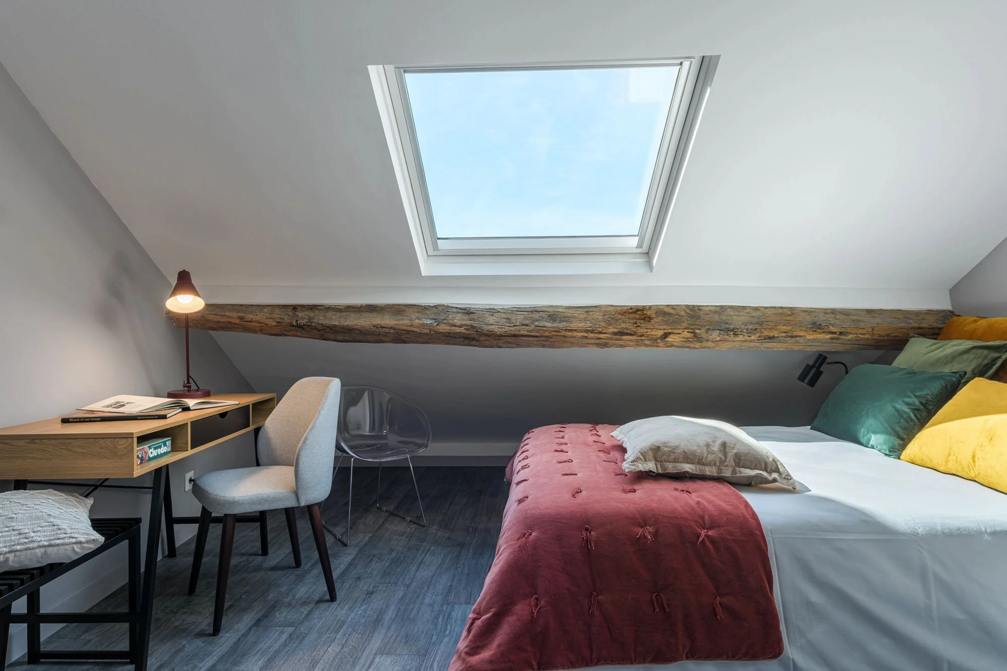 Chambre sous un toit mansardé avec plafond en pente, lit avec oreillers de couleurs contrastantes, grande fenêtre de toit laissant entrer la lumière naturelle, bureau avec lampe de lecture, chaises transparentes et sol en bois foncé.