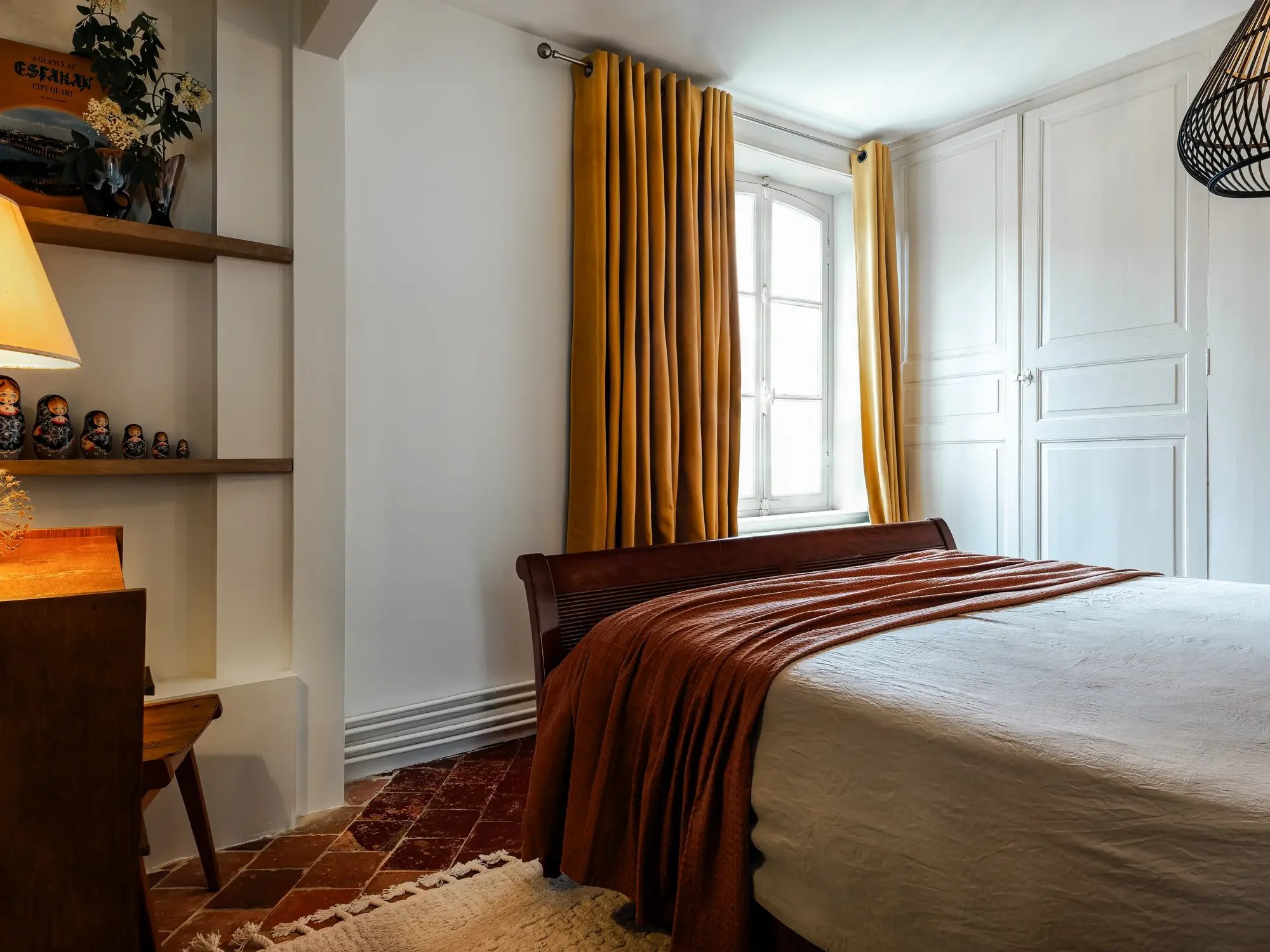 Chambre avec lit, fenêtres à volets, rideaux jaunes, étagères avec décoration et lampadaire