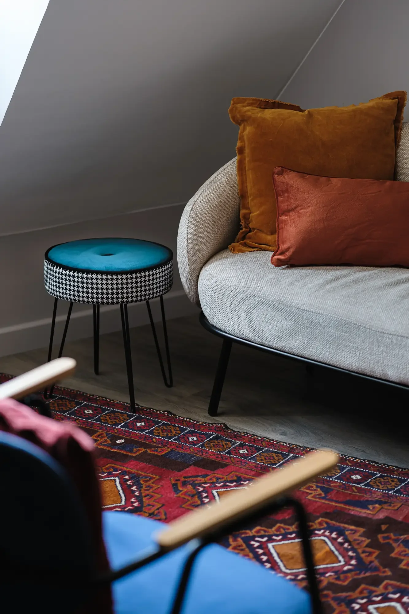 Coin salon avec un canapé beige, deux coussins orange et marron, un tapis oriental rouge et bleu, une petite table à hiboux à motifs pied-noir et un mur gris incline.