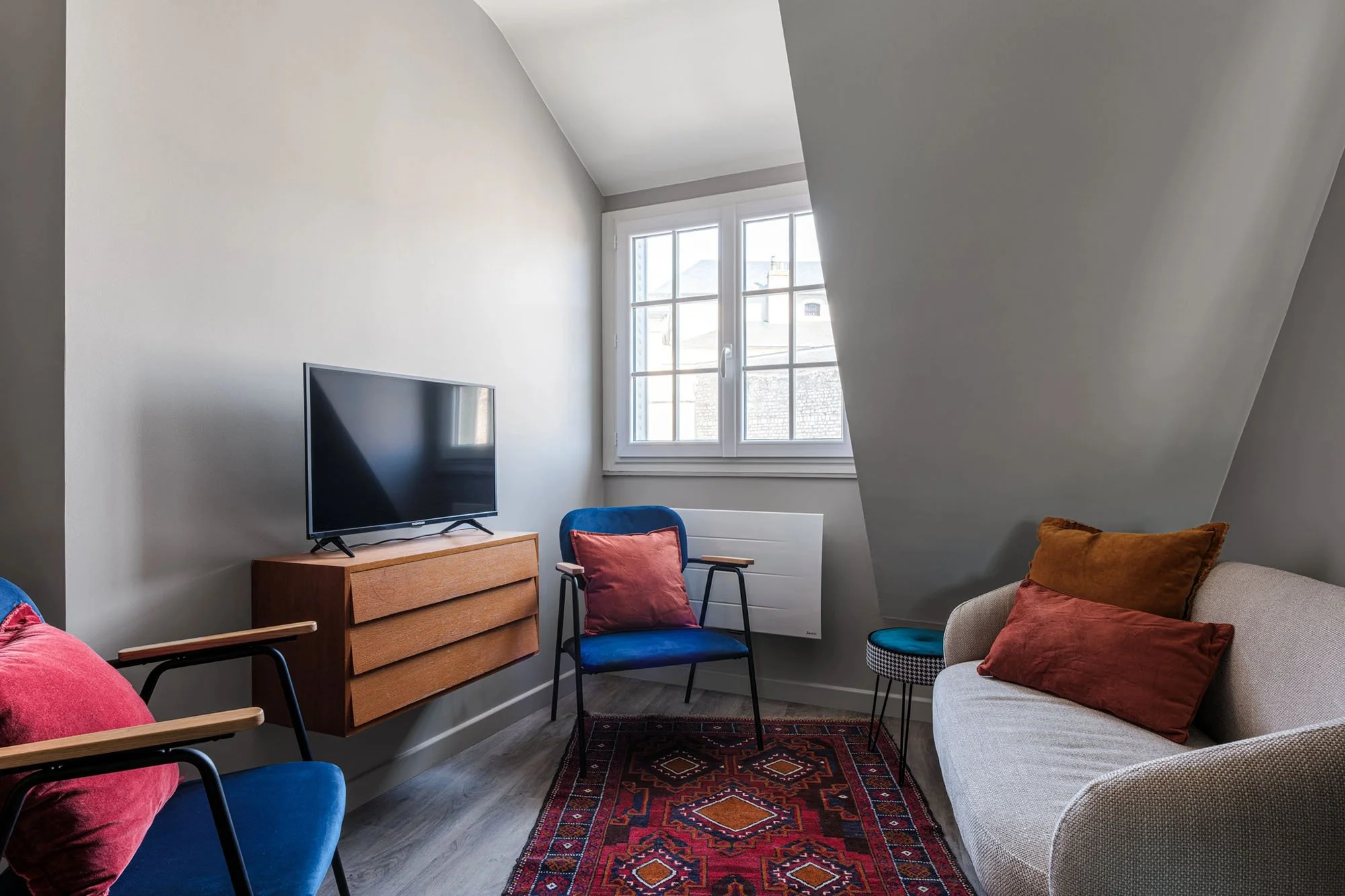 Salon avec fauteuils bleus, sofa beige, tapis berbère, télévision sur meuble en bois, fenêtres avec vue sur des bâtiments.