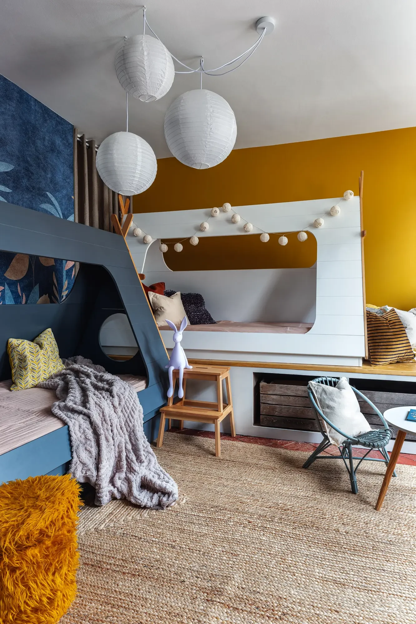 Une chambre pour enfants avec un lit en forme de maison blanche, décorée de guirlandes lumineuses, un lit en forme de cabane bleue, coussins et couvertures, une chaise en rotin avec un coussin blanc, un tapis en jute, une lampe en papier blanche, et 