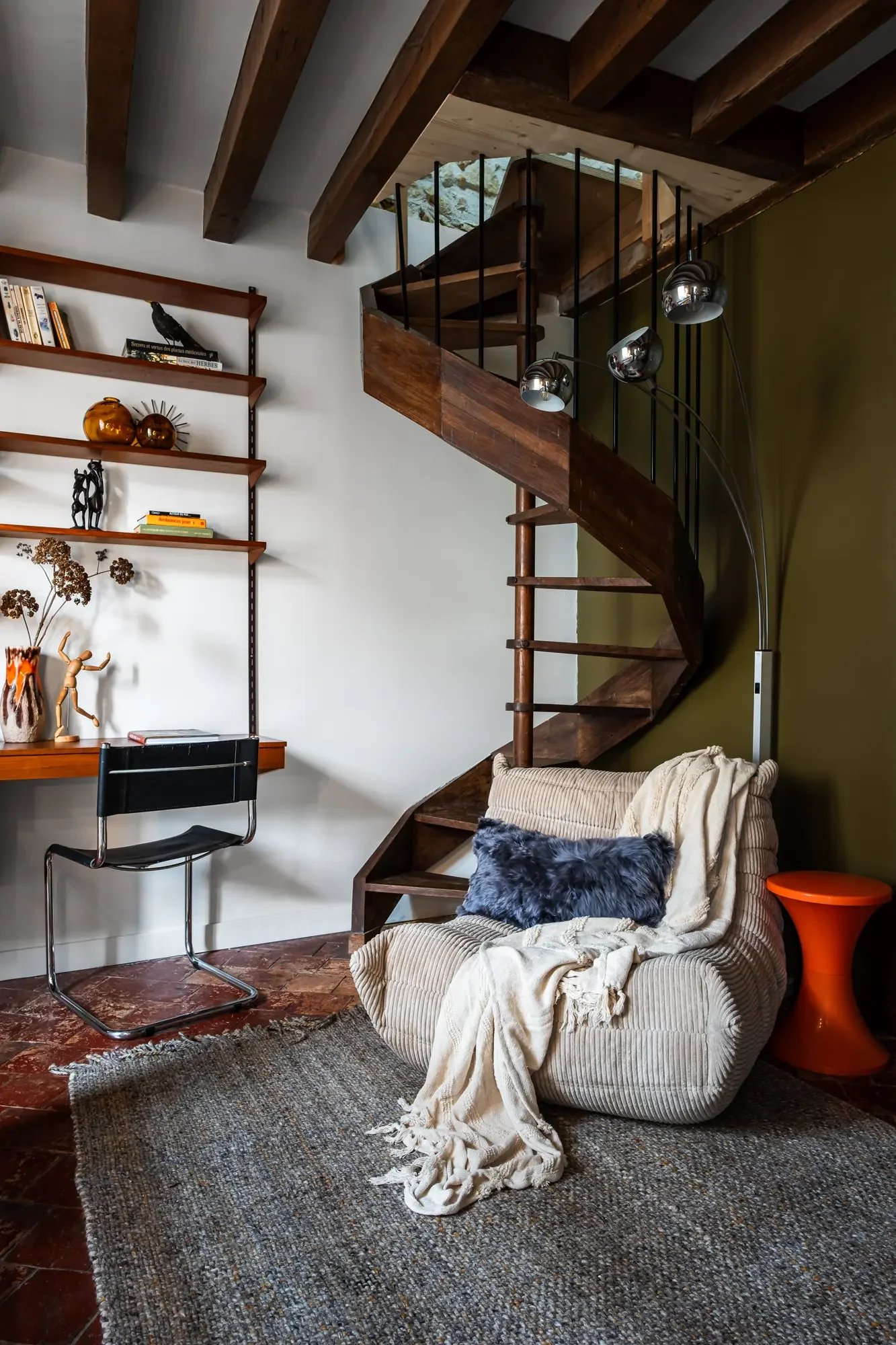 Intérieur d'un salon avec un escalier en bois, une chaise, une étagère avec des livres et des décorations, un fauteuil avec un coussin en fourrure, une lampe d'angle, et un tapis au sol.