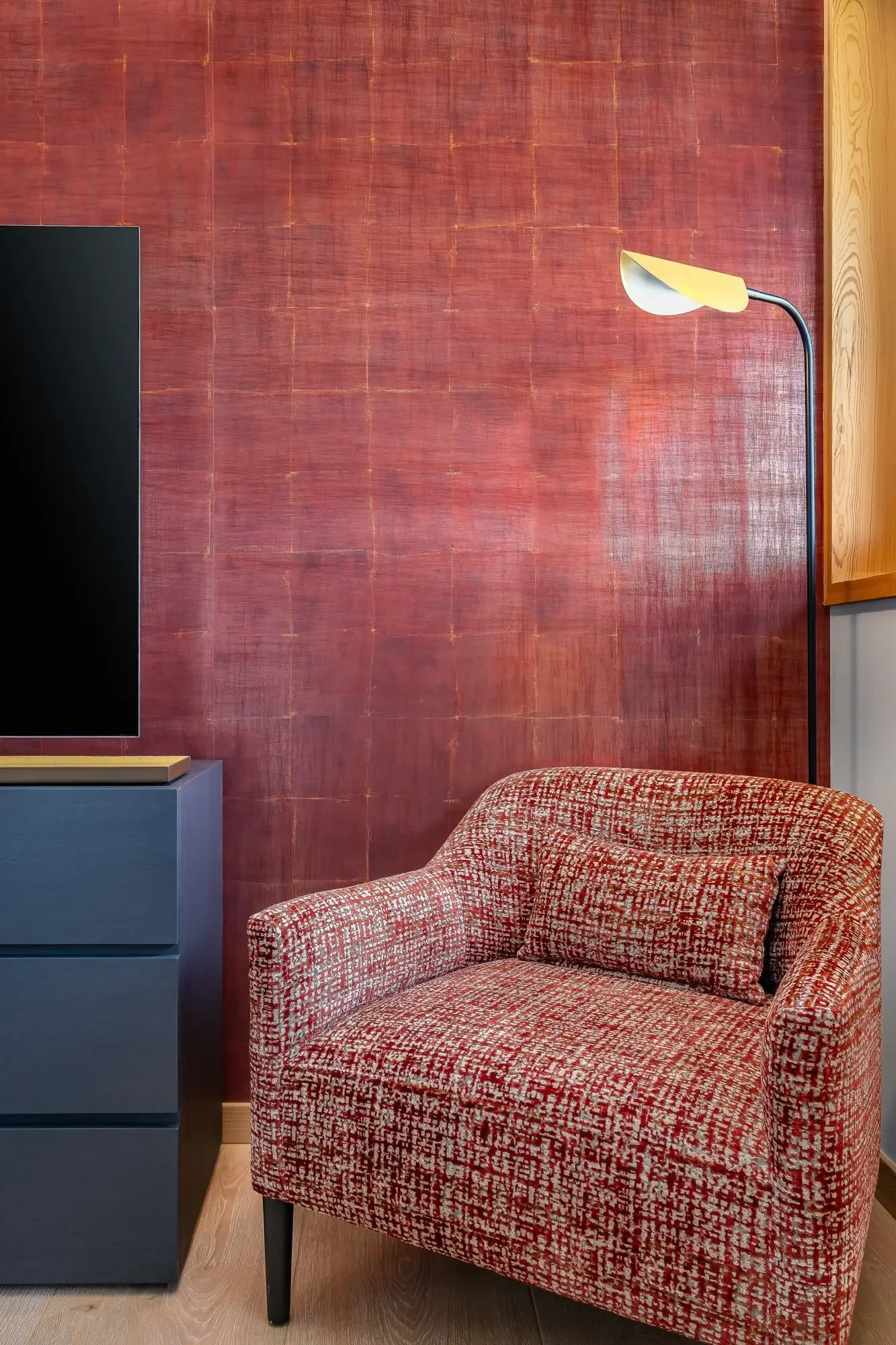 Un fauteuil rouge et beige avec un coussin, à côté d'une commode bleue, devant un mur rougeâtre et une lampe au mur.