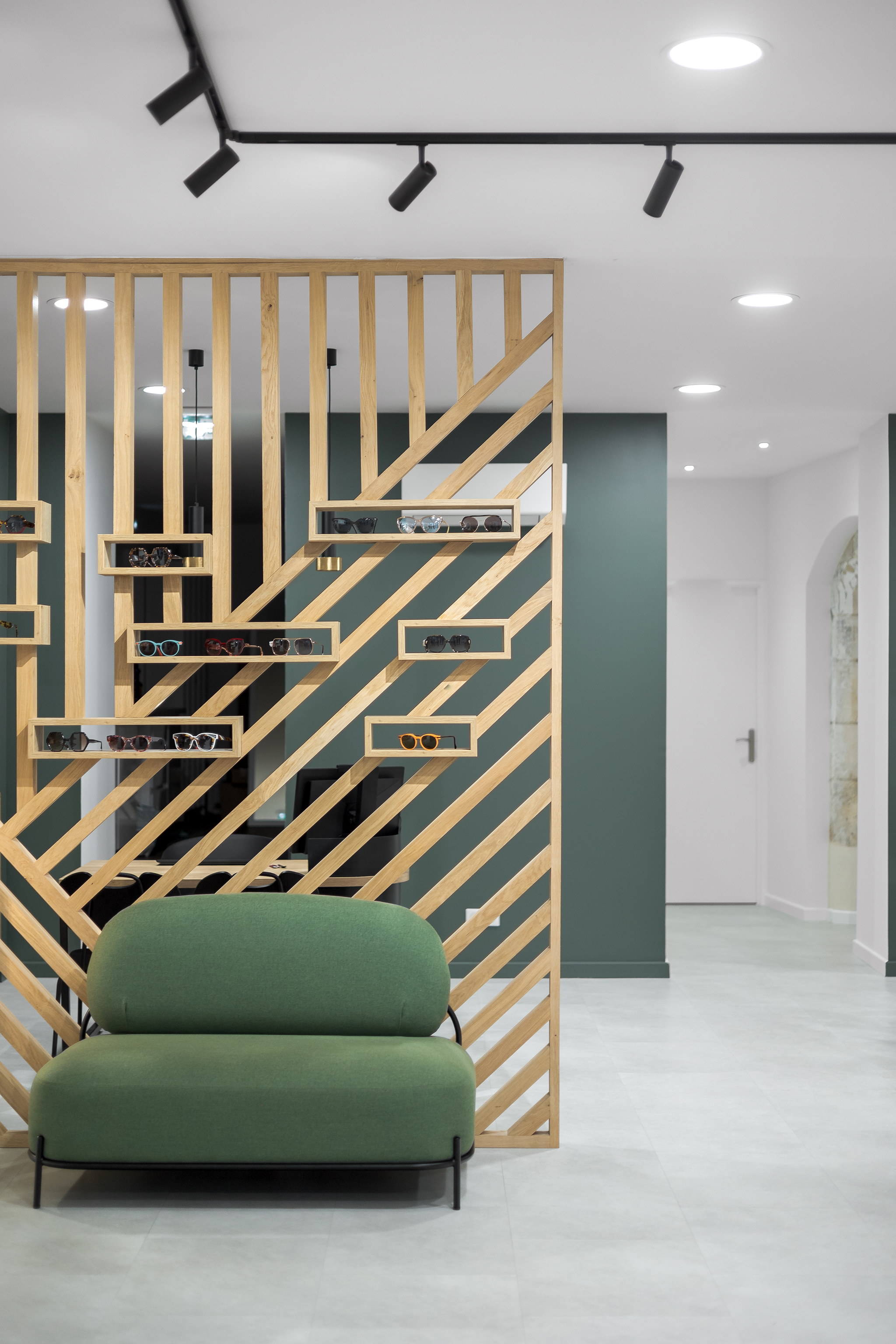 Espace intérieur moderne avec un mur en bois ajouré affichant des lunettes de soleil, un canapé vert en premier plan, et un plafond avec des spots lumineux.