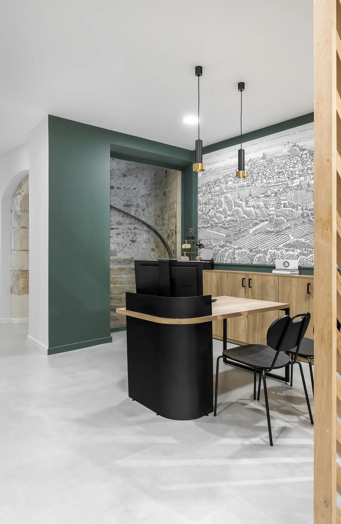 Bureau moderne avec deux ordinateurs, chaise en bois, mur avec fresque de ville en noir et blanc, suspension design, et mur en pierre visible à travers une ouverture.