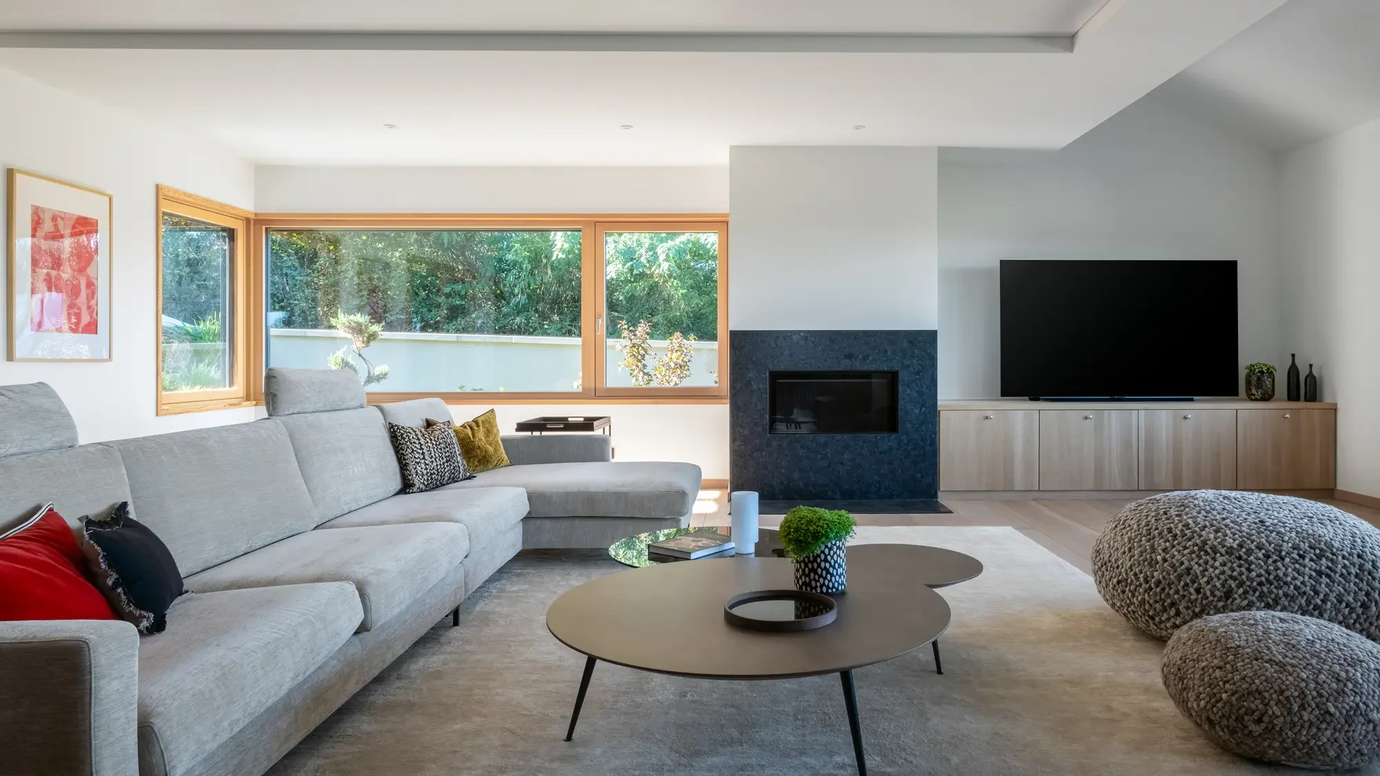 Salon moderne avec canapé gris, table basse, grands poufs gris, cheminée, télévision sur meuble en bois, grande fenêtre laissant entrer la lumière naturelle, décor minimaliste.