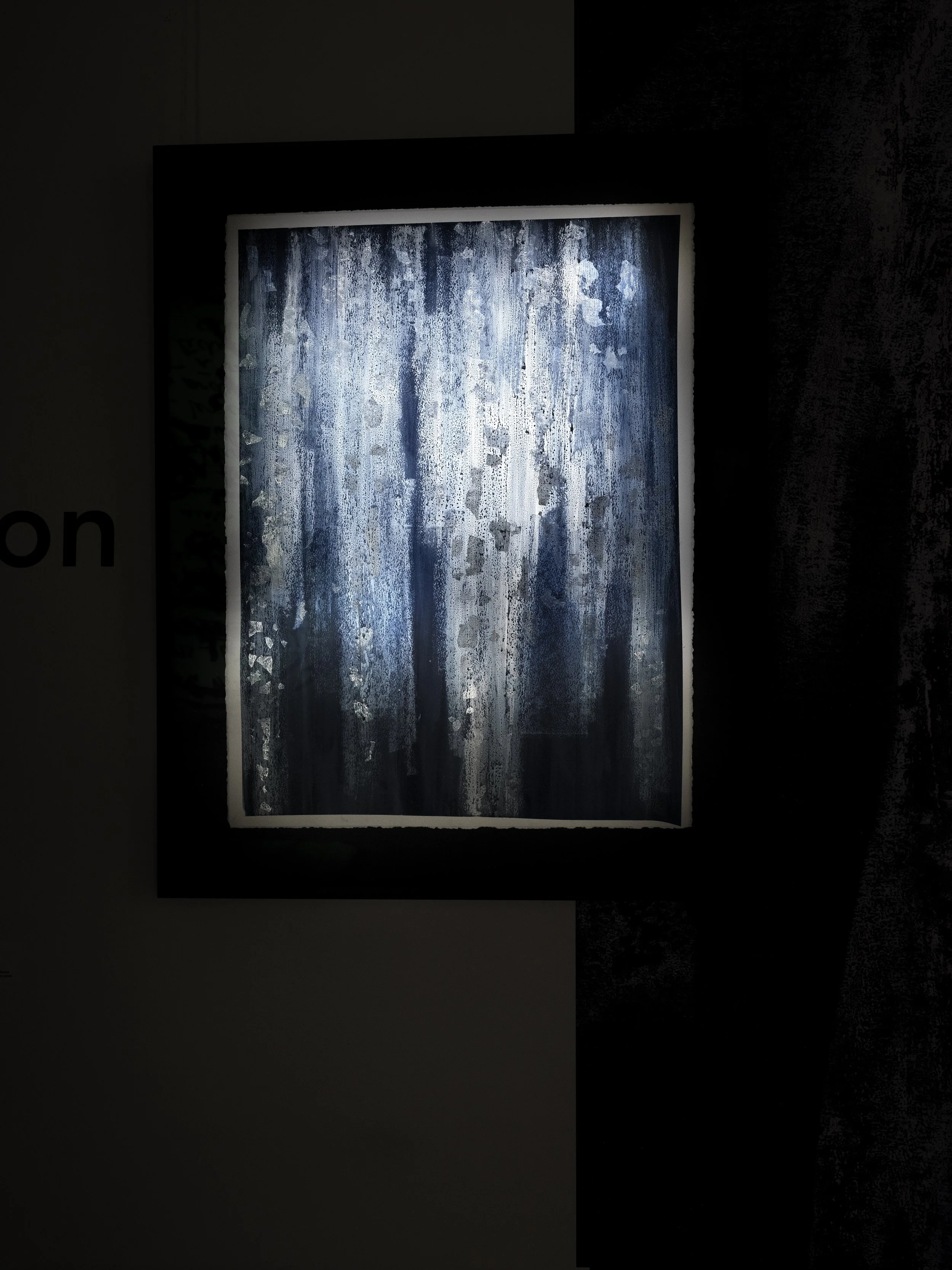 Une peinture abstraite avec des tons de bleu, blanc et gris, encadrée et éclairée dans une pièce sombre.