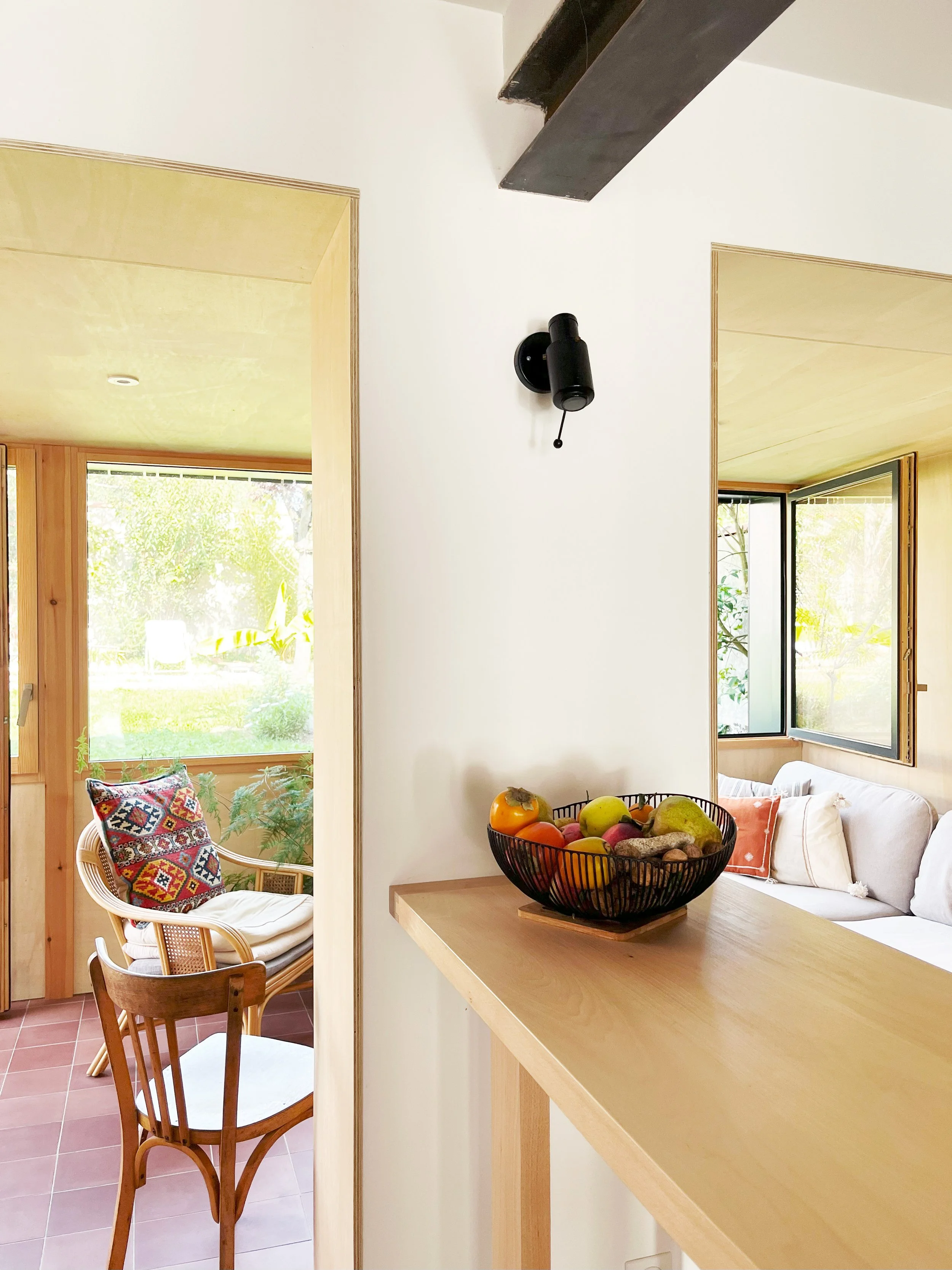 Intérieur d'une maison avec un coin salon lumineux, fenêtre ouverte, un panier de fruits sur un comptoir en bois, et des meubles en bois avec coussins.