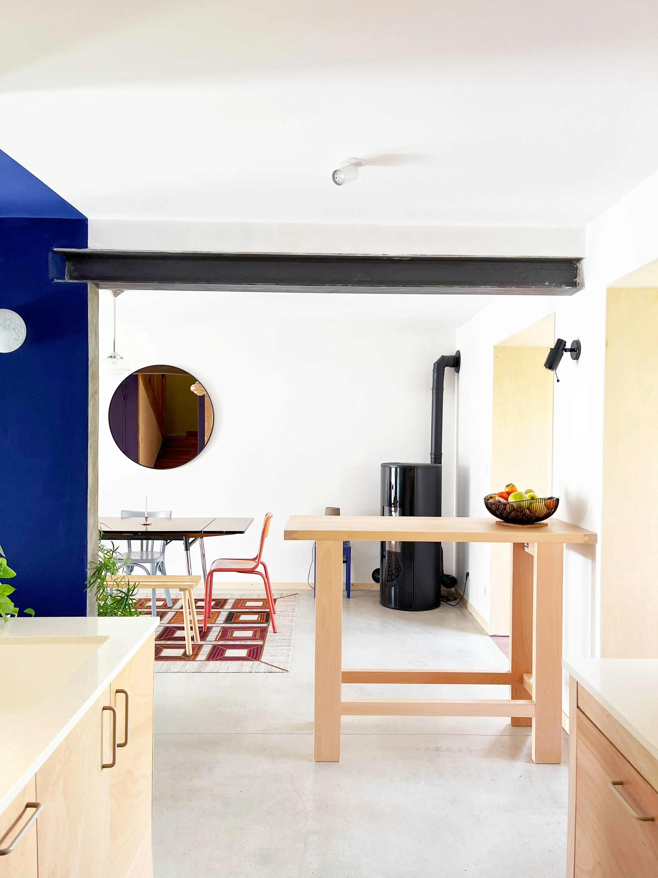 Intérieur moderne avec cuisine, table à manger, chaises colorées, miroir rond et poële à bois.
