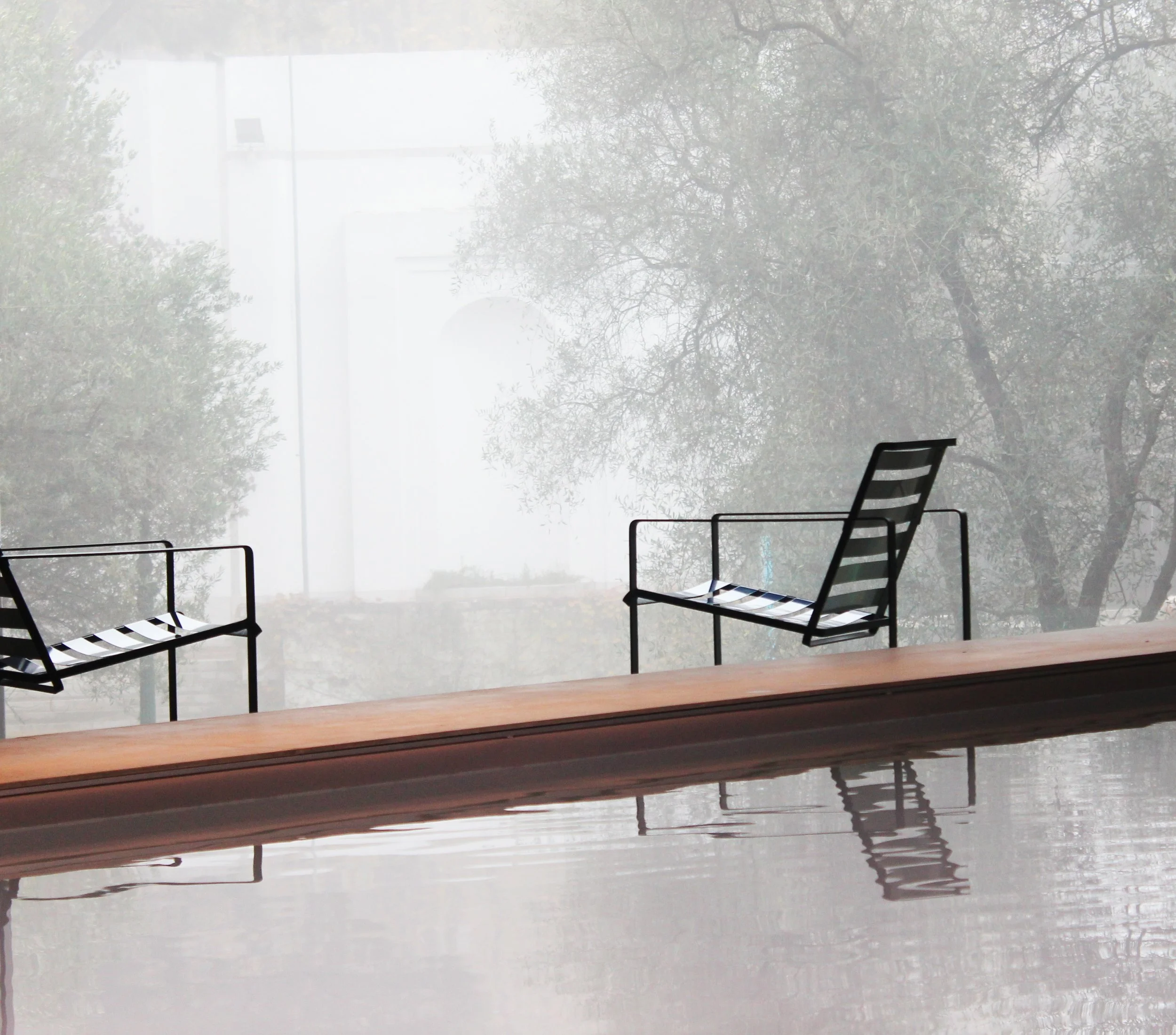 Chaises en métal et bois rouge autour d'une piscine calme, vue à travers une fenêtre floue avec brouillard et arbres en arrière-plan.