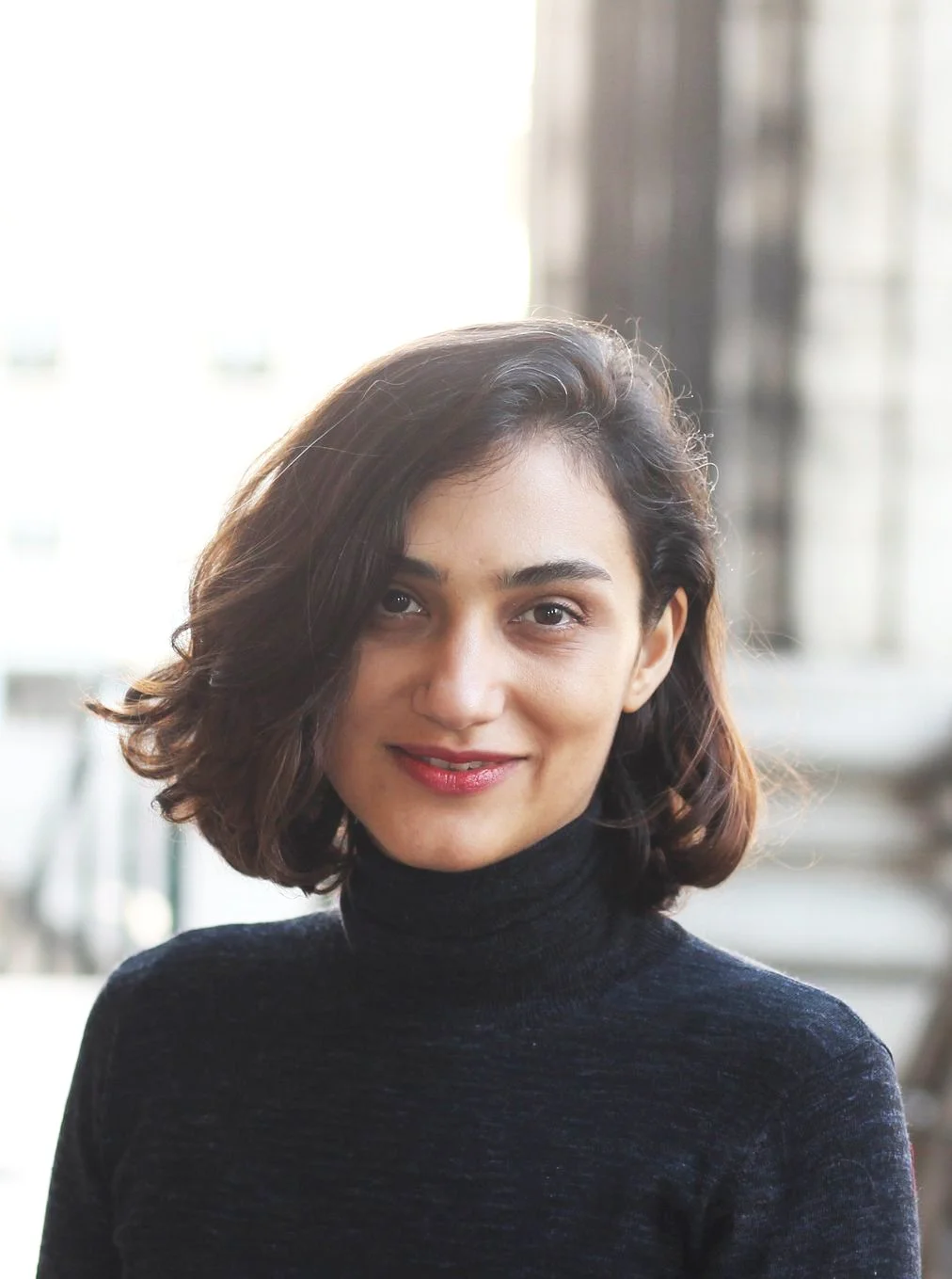 Portrait d'une femme avec des cheveux bruns, souriant, portant un pull noir à col roulé, fond flou d'un intérieur lumineux.