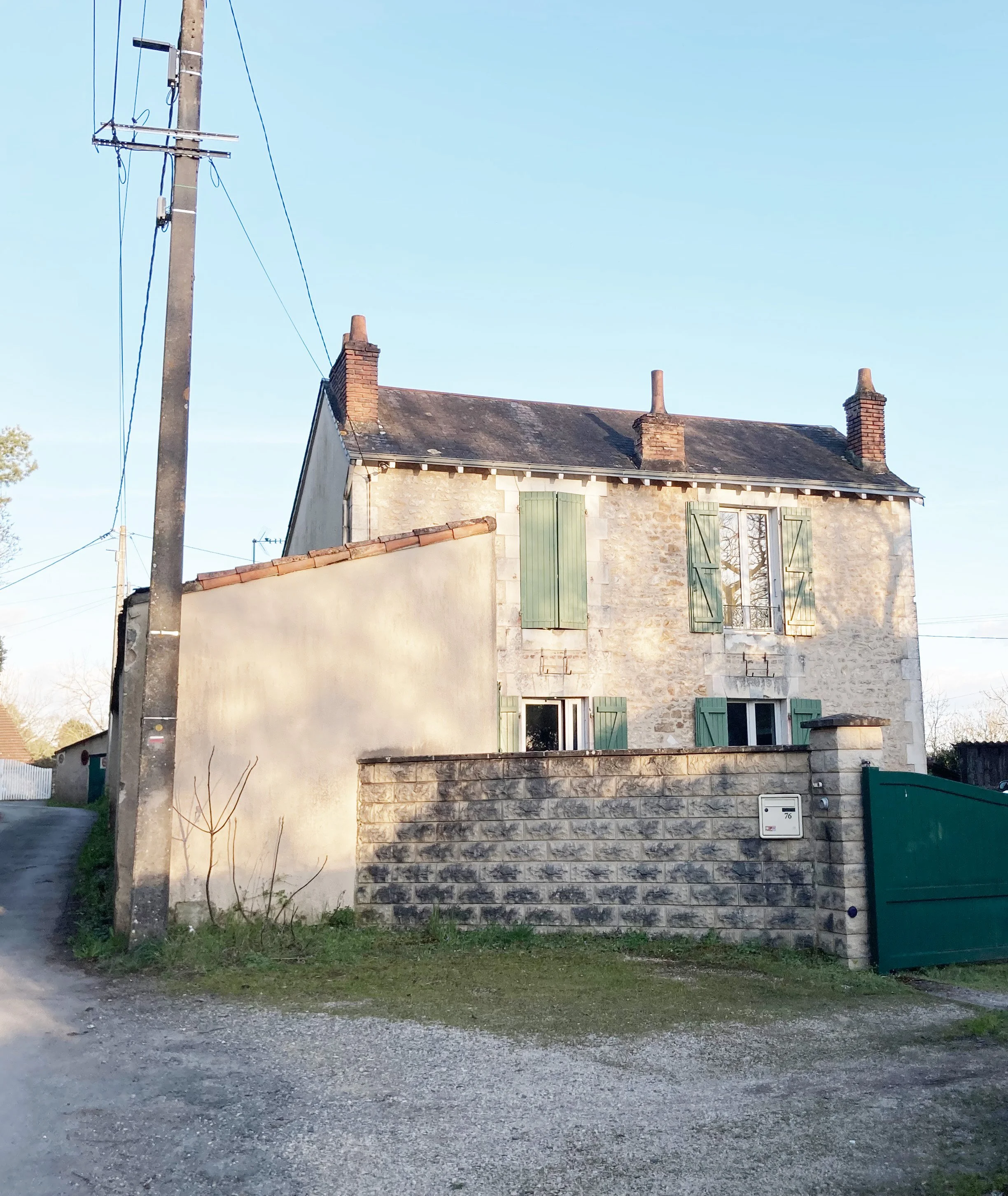 Maison en pierre avec volets verts, mur en pierre, clôture en pierre et porte de garage verte, sous un ciel clair.