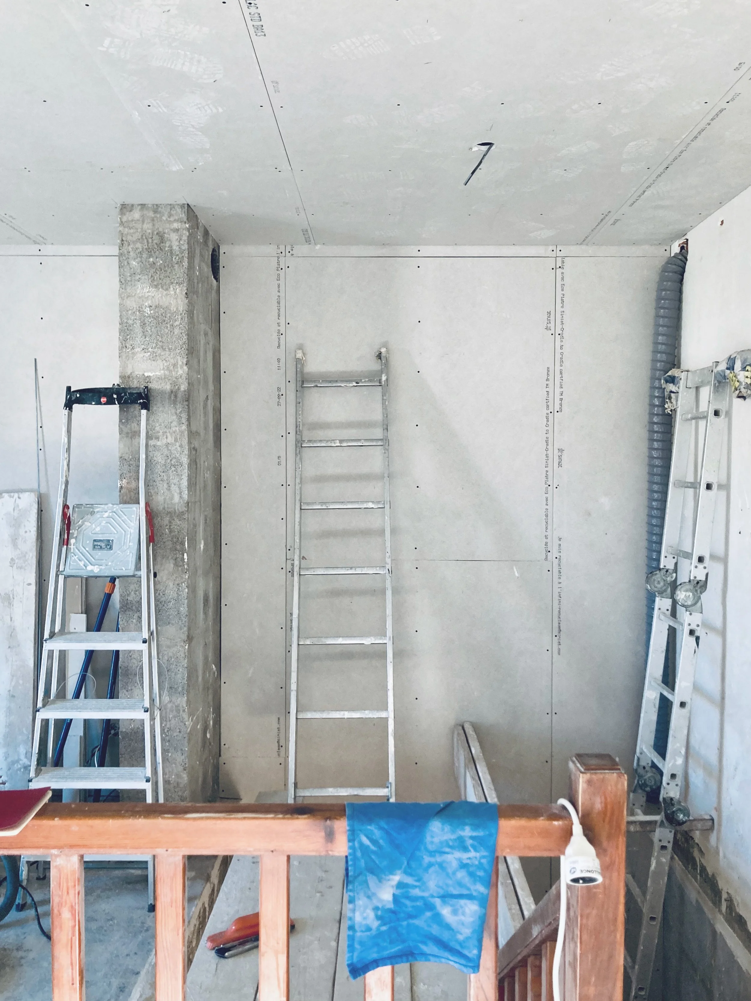 Pièce en cours de rénovation avec murs en plâtre, échelles, un tuyau flexible, une lampette électrique, et un linge bleu suspendu sur une barrière en bois.