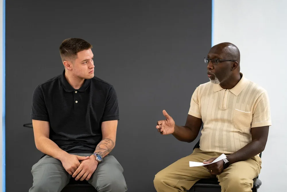 Two men are engaged in a conversation, sitting on chairs against a plain background. The man on the left is young, with short brown hair, wearing a black polo shirt and gray pants. The man on the right is older, bald, with glasses, wearing a beige polo shirt and tan pants, holding papers in his hand.