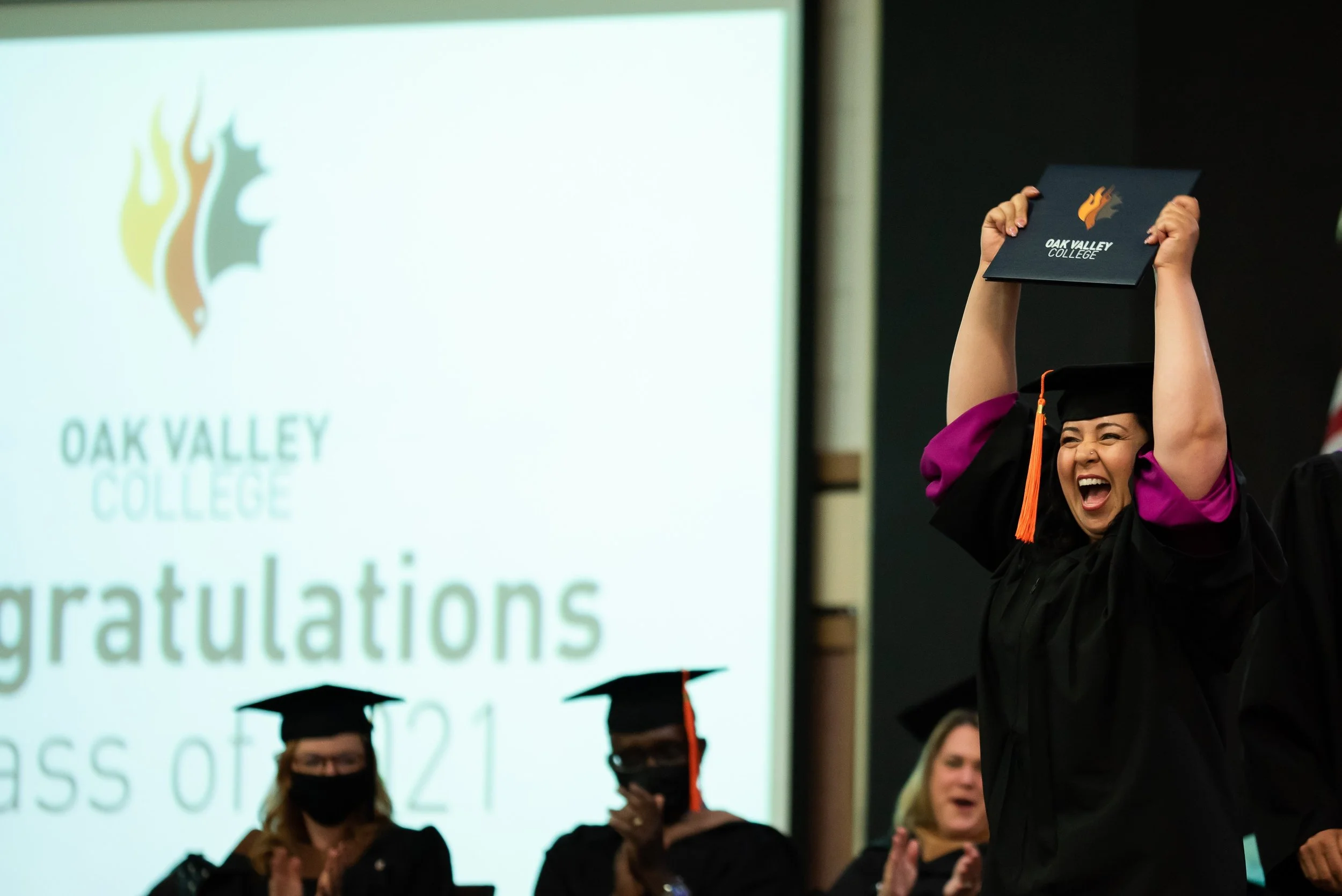 Happy graduate woman in cap and gown celebrating with a diploma at graduation ceremony, with other graduates and a large screen displaying Oak Valley College logo and congratulatory message in the background.