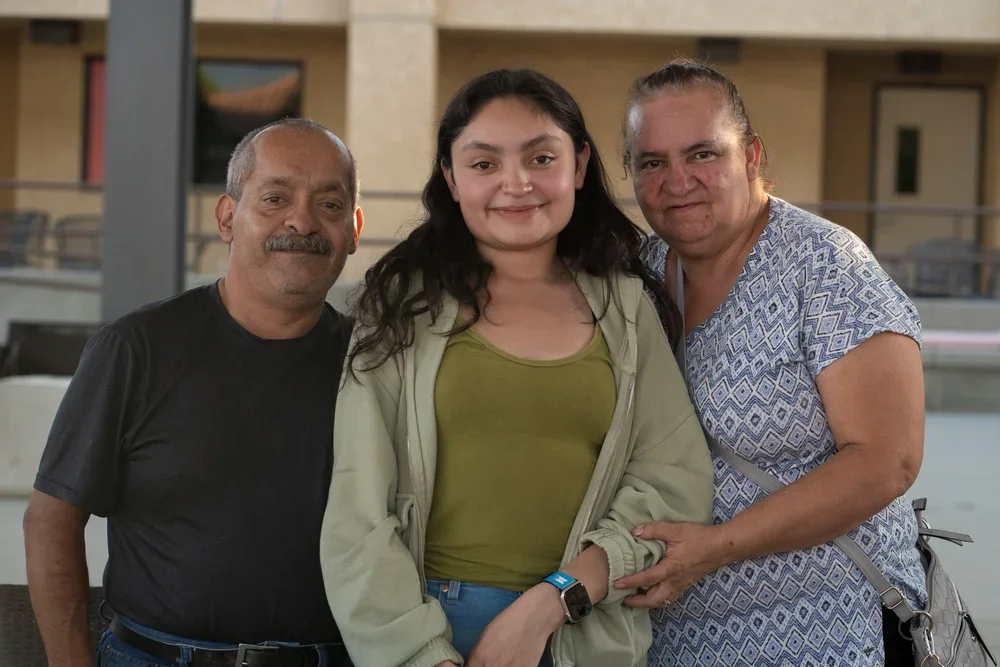 Three people, a man, a young girl, and a woman, standing close together and smiling indoors.