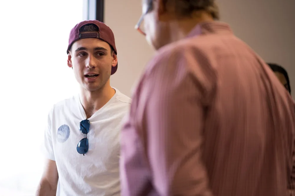 A young man wearing a backward hat and sunglasses around his neck is speaking to an older man with glasses and a pink striped shirt in an indoor setting.