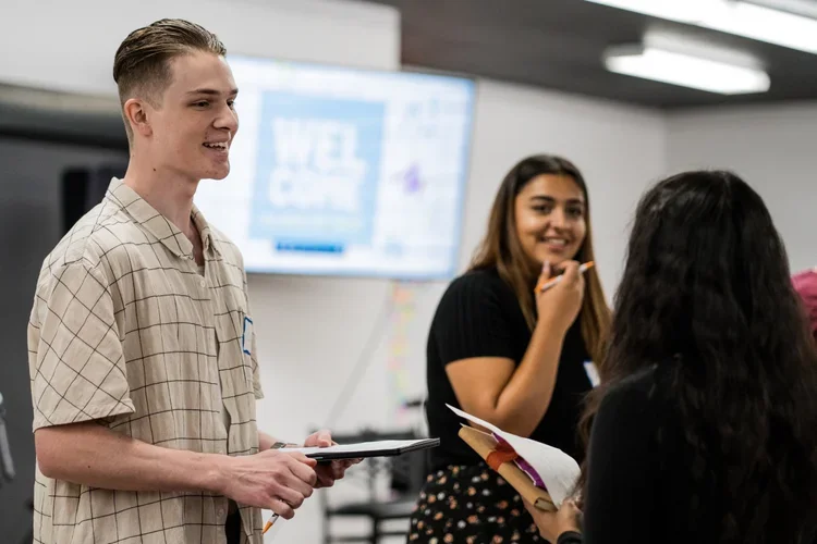 Young man and young woman in a classroom or office setting, engaged in conversation or a discussion, with a blurred presentation screen in the background.