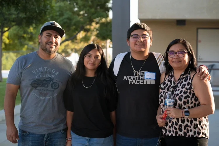 Group of four people standing outdoors, smiling, with trees and a building in the background.