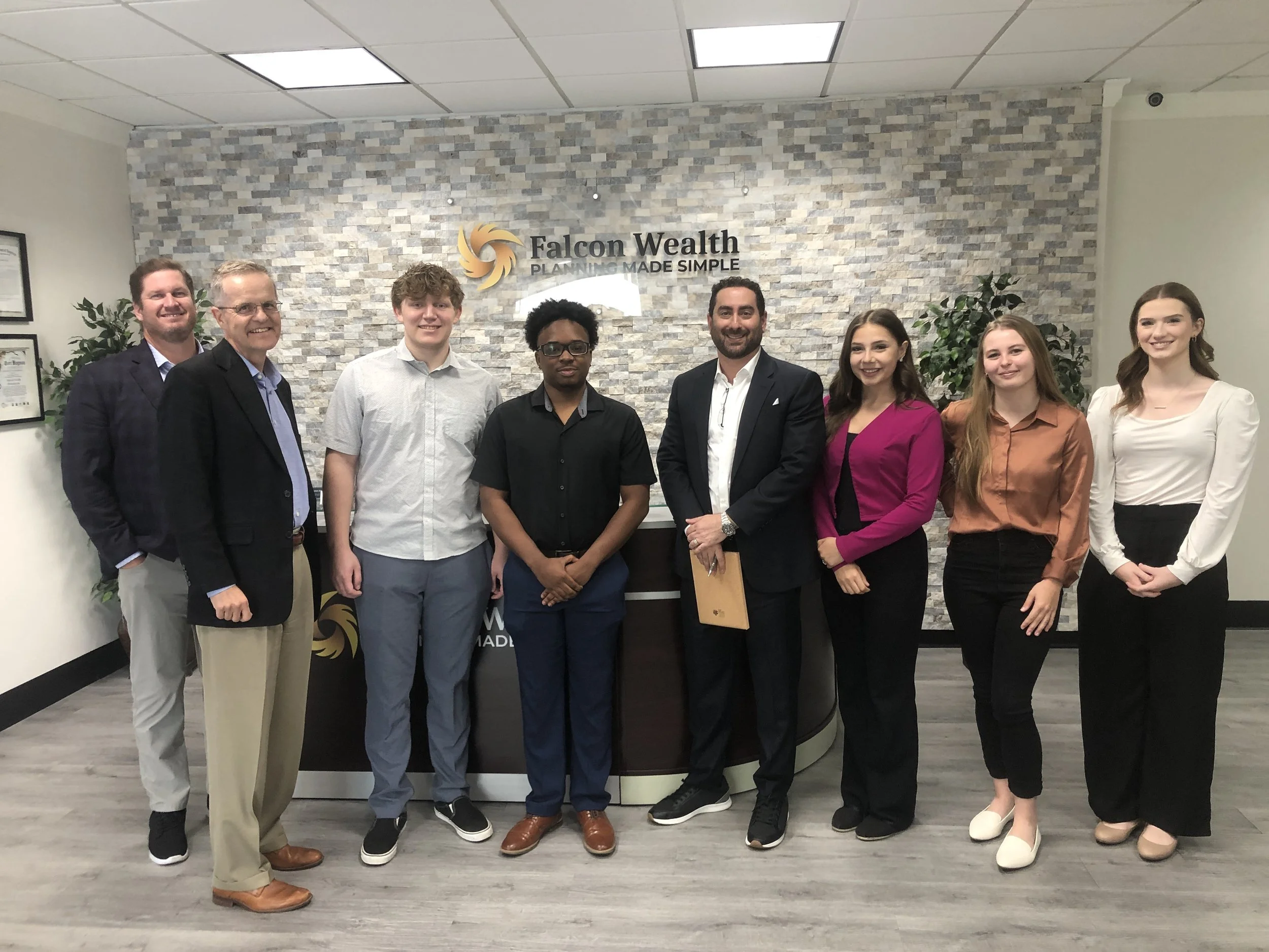 Group of nine people standing in a row in an office with a stone wall background, Falcon Wealth sign, and plants.
