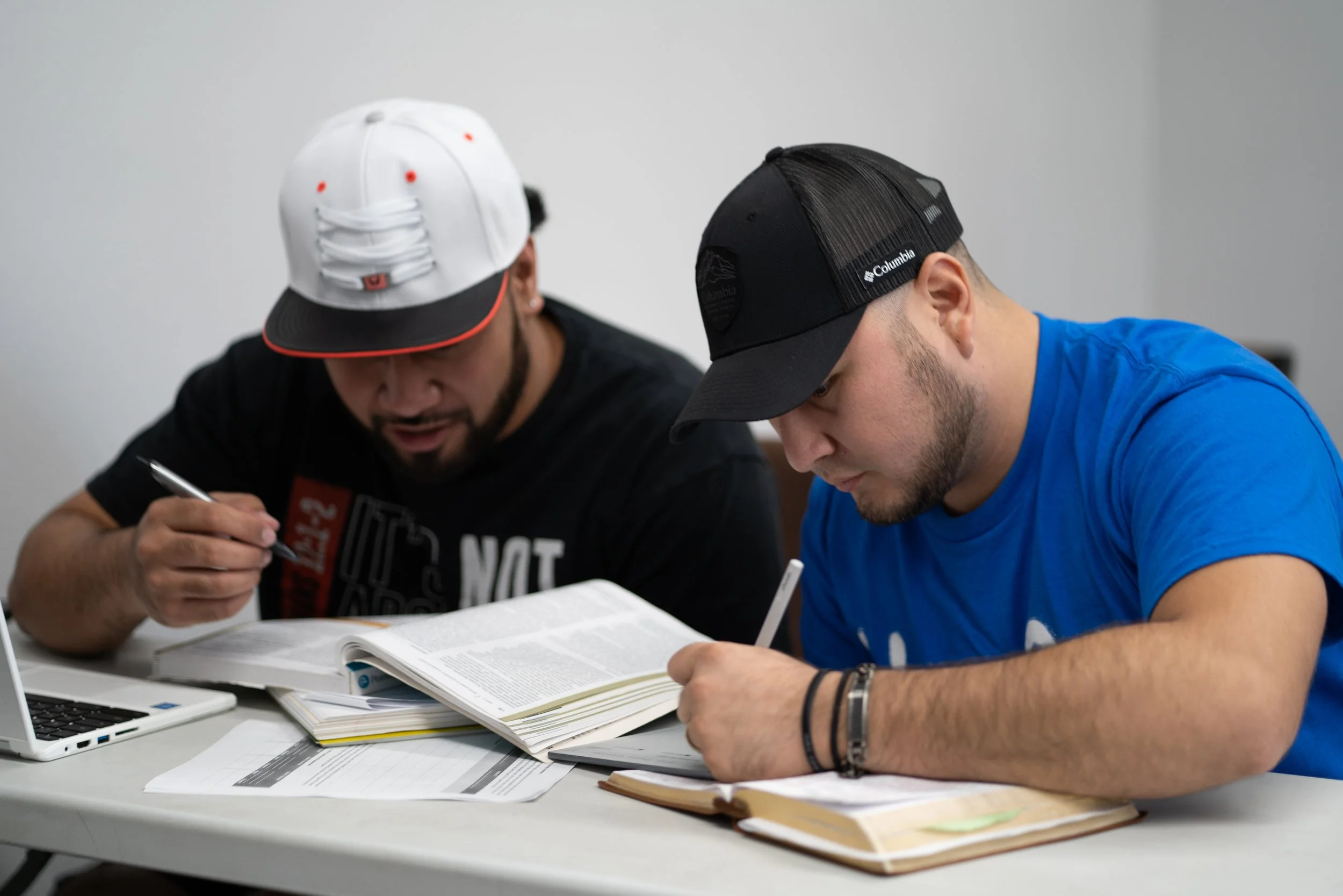 Two young men studying together at a table with open textbooks, notebooks, and a laptop in front of them.