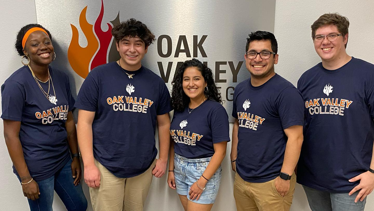 Group of five diverse young adults standing together, smiling, in front of a sign that reads 'OAK VALLEY',