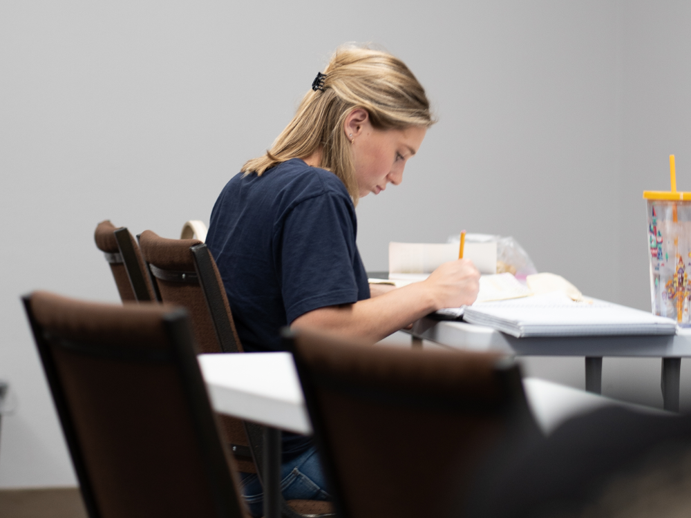 A young woman with blonde hair sitting at a table, writing in a notebook with a pencil. There are binders and notebooks on the table, and a drink with a colorful straw.