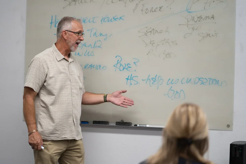 A middle-aged man with gray hair, glasses, and a beard stands facing a whiteboard, gesturing with his right hand. There is a woman with blonde hair sitting in front of him, listening. The whiteboard behind the man has handwritten notes in blue and black ink.