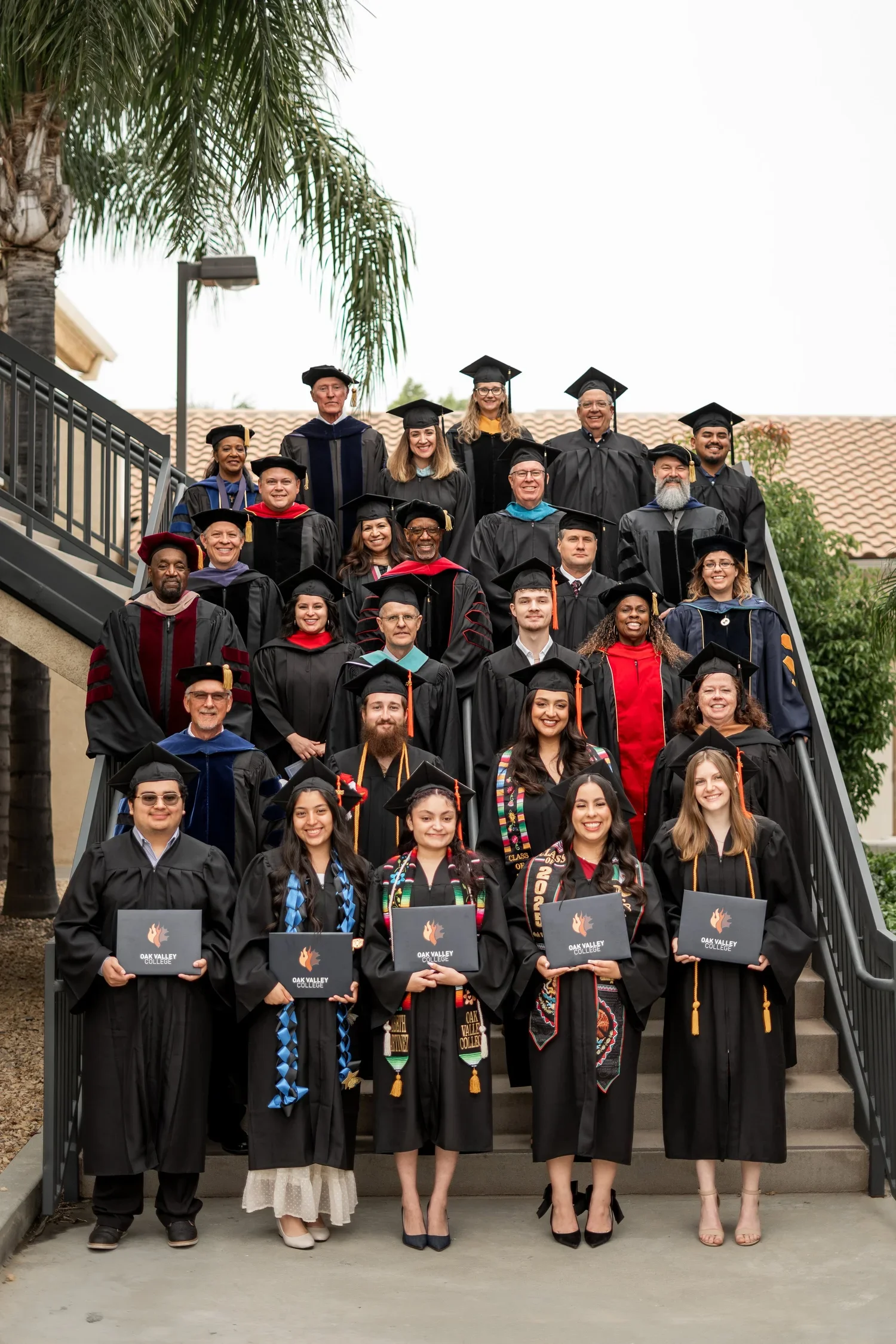 Group of graduates in caps and gowns on outdoor staircase, some holding diplomas, at Oak Valley College.