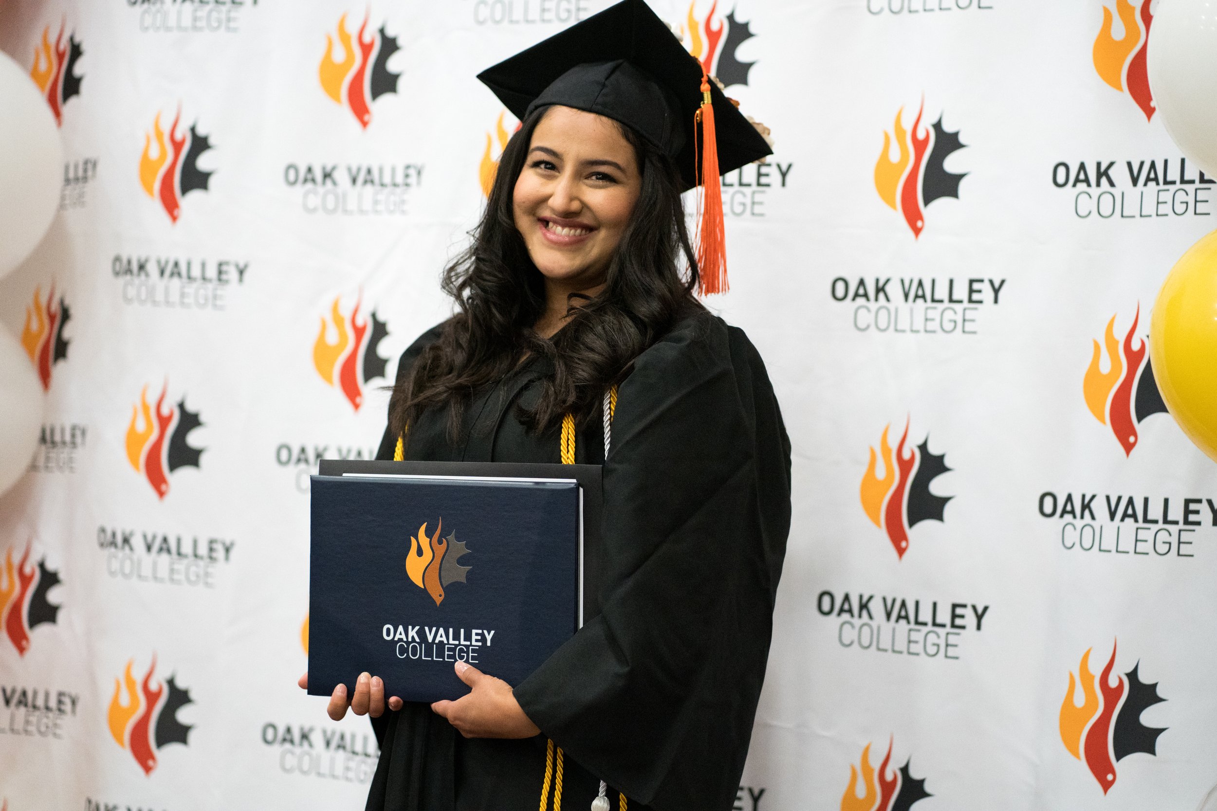 A young woman in graduation cap and gown holding a diploma in front of a backdrop with Oak Valley College logo, smiling at the camera.