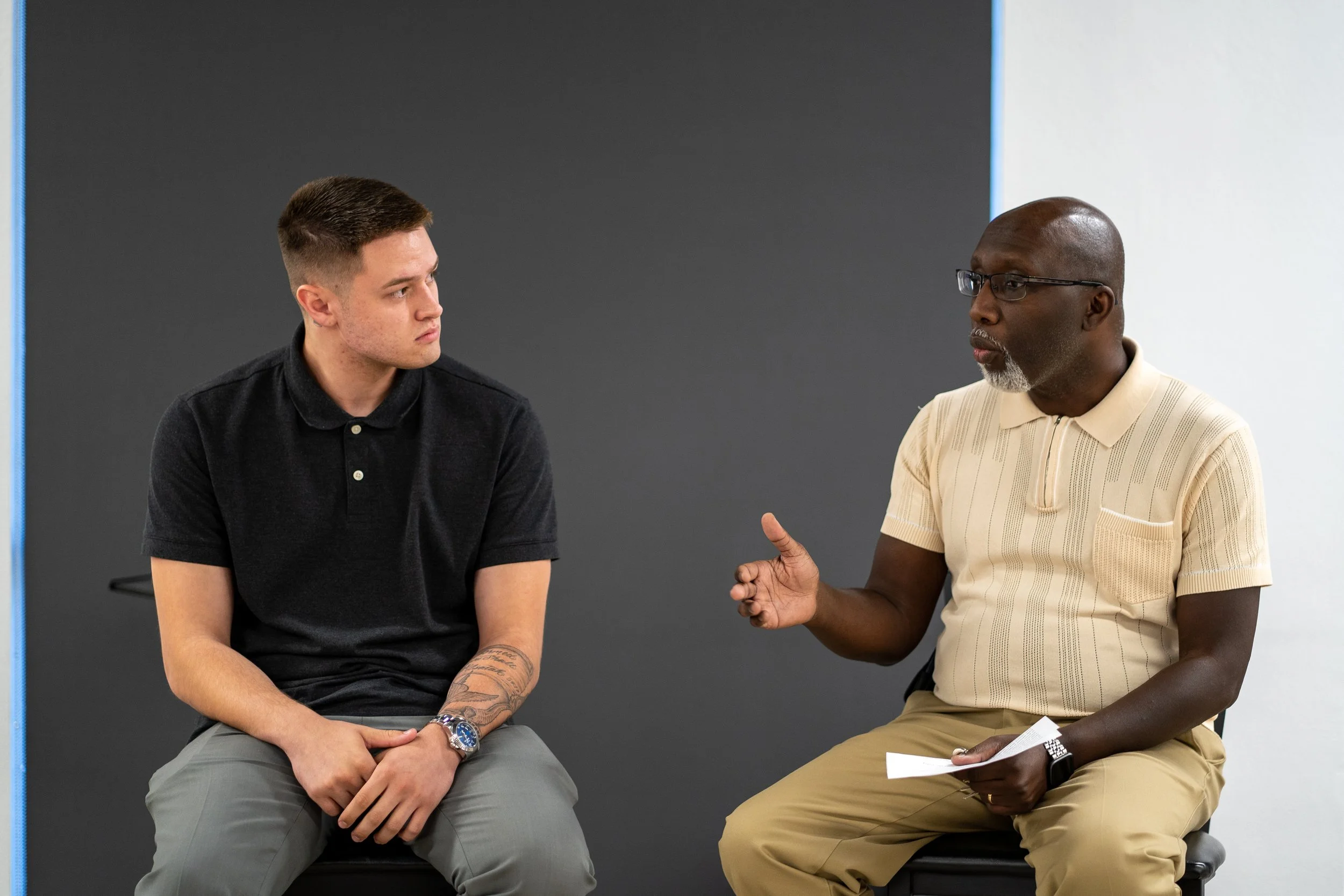 A young man in a black polo shirt and gray pants sitting and listening to an older man in a beige polo shirt with glasses, who is speaking and gesturing with his right hand, holding a paper in his left hand. They are seated against a plain, dark and light gray background.