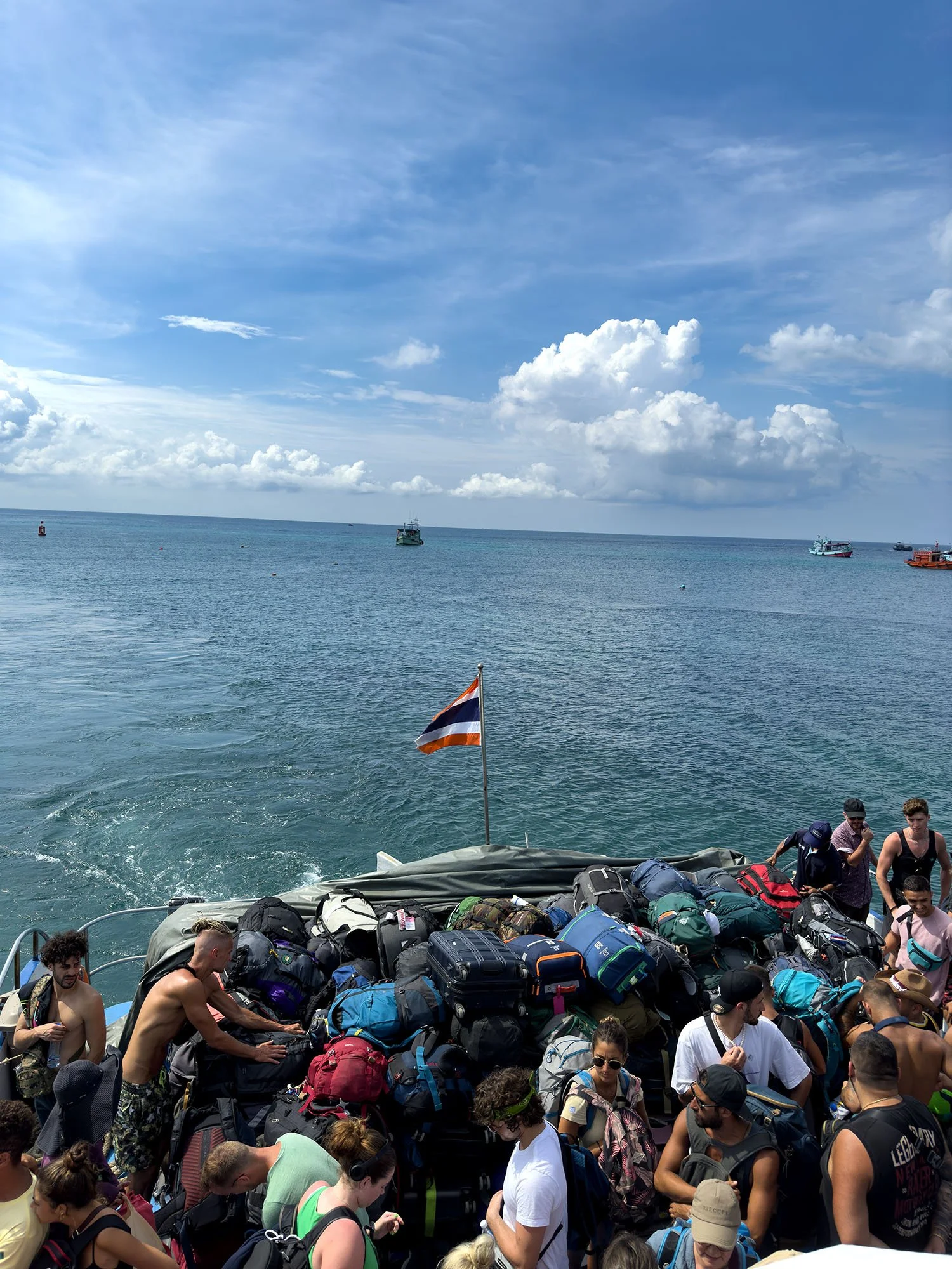 Crowd of people with backpacks on a boat at sea, with a flag, ocean in the background, and several other boats in the distance.