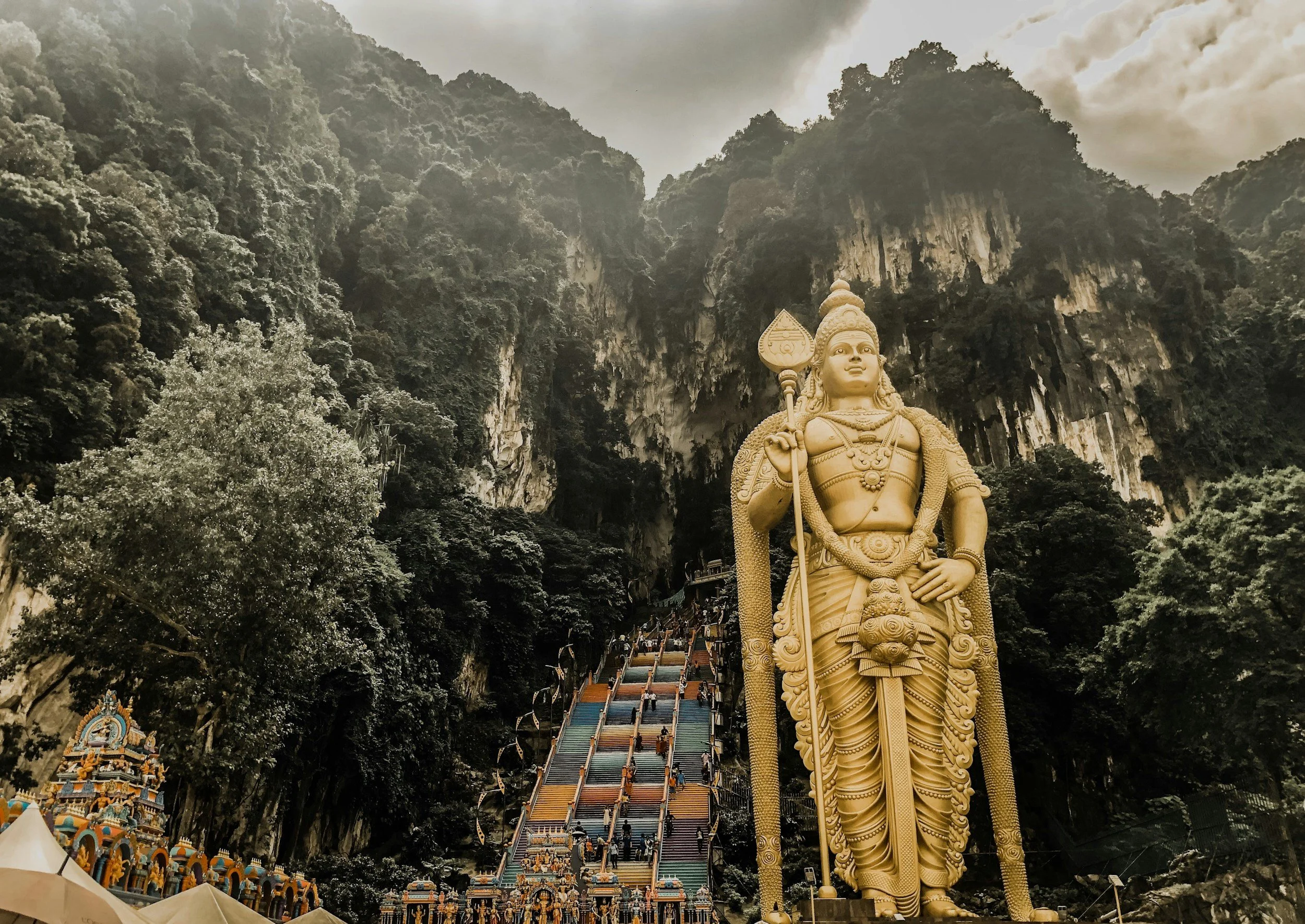Grote gouden standbeelden van een hindoeïstische godheid bij de Batu Caves, met een kleurrijk trappenpad en een grottenrots op de achtergrond.