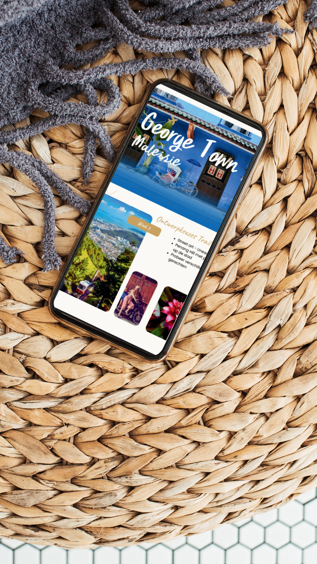 Een smartphone ligt op een gevlochten rieten oppervlak. Op het scherm is een website te zien met de titel 'George Town' en foto's van een stad, een landschap, en bloemen.