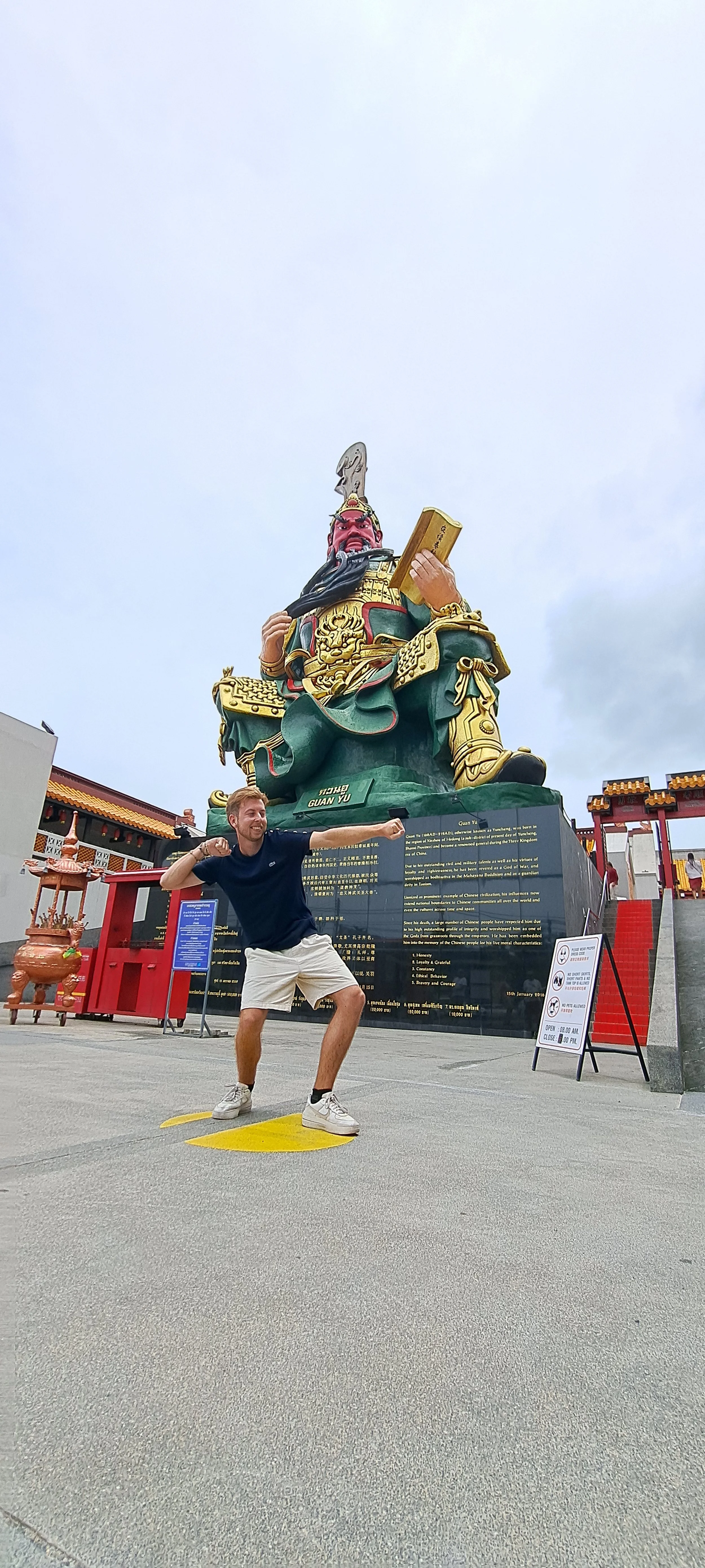 Man poseert voor een grote Chinese godenbeeld, gekleed in casual kledij, buiten onder een bewolkte hemel.