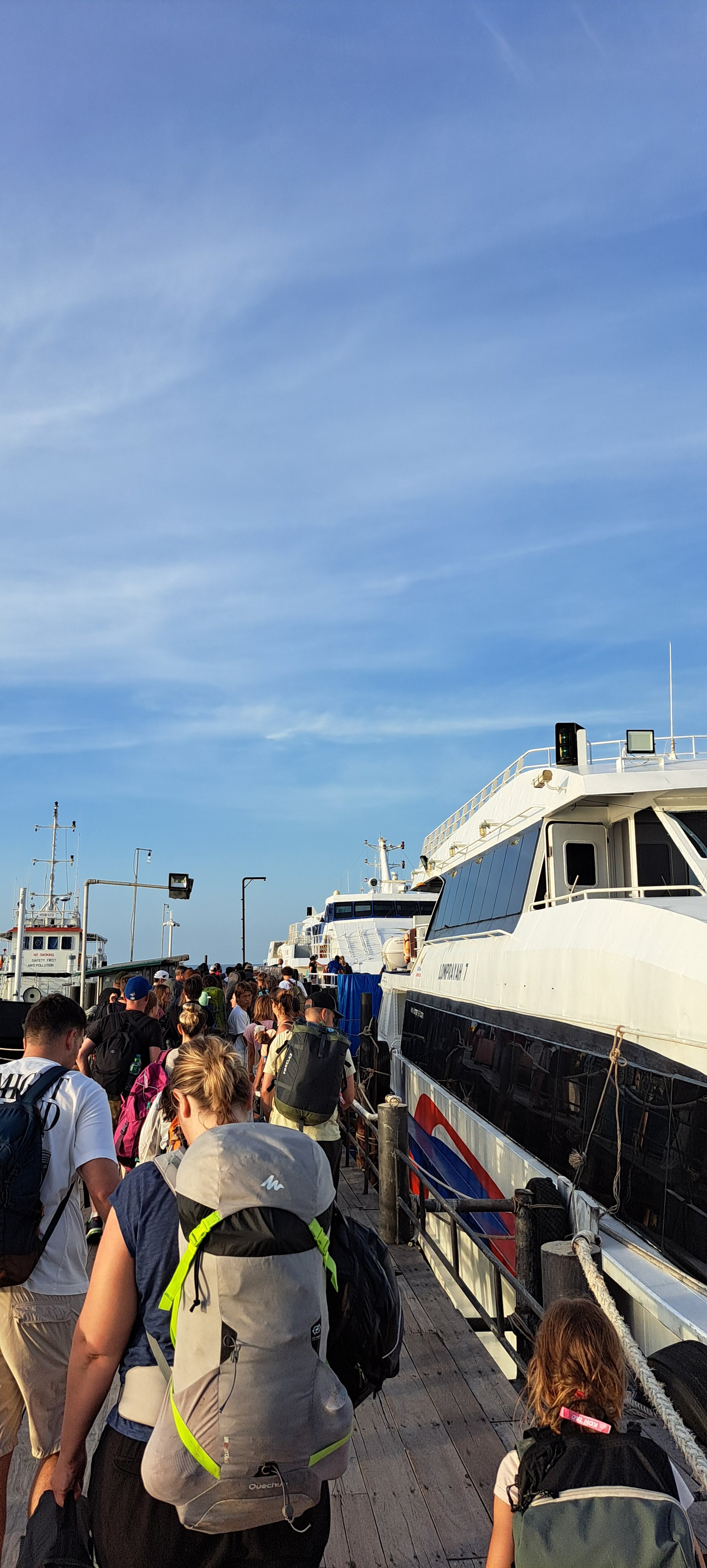 Veel mensen staan in de rij op de pier, klaar om de boot te nemen, met verschillende kleuren backpacks en kleding, naast een passagiersschip met een blauwe en witte romp, onder een heldere blauwe hemel.