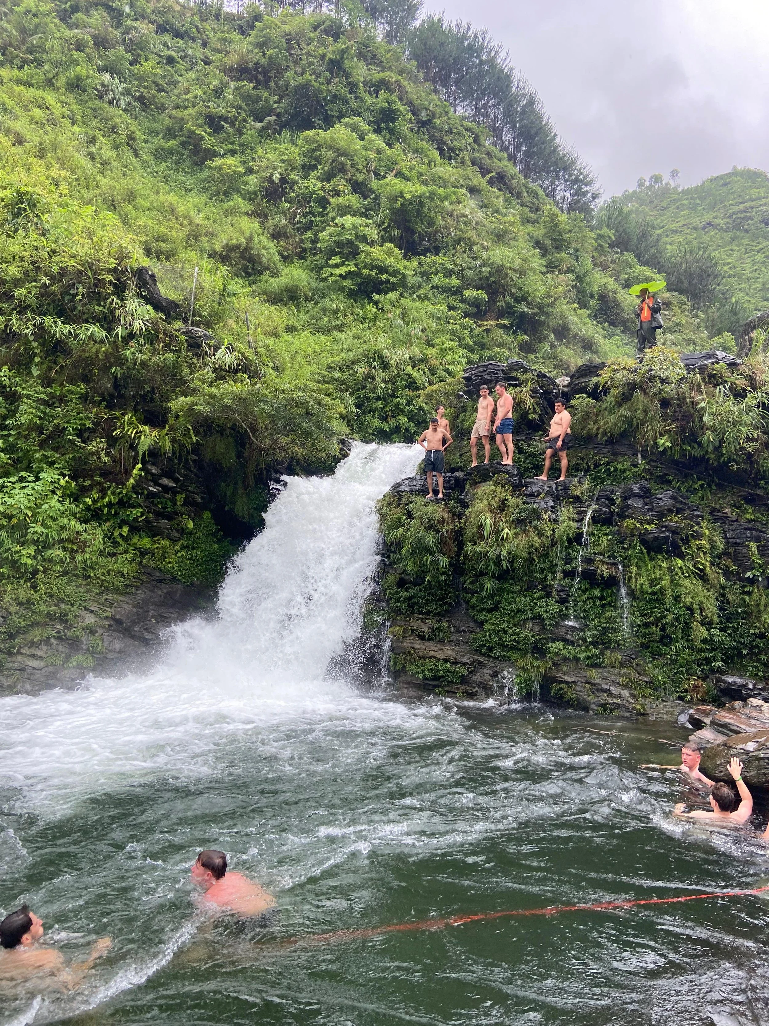 Mensen zwemmen in een natuurlijke rivier met waterval, omringd door groene vegetatie en heuvels.