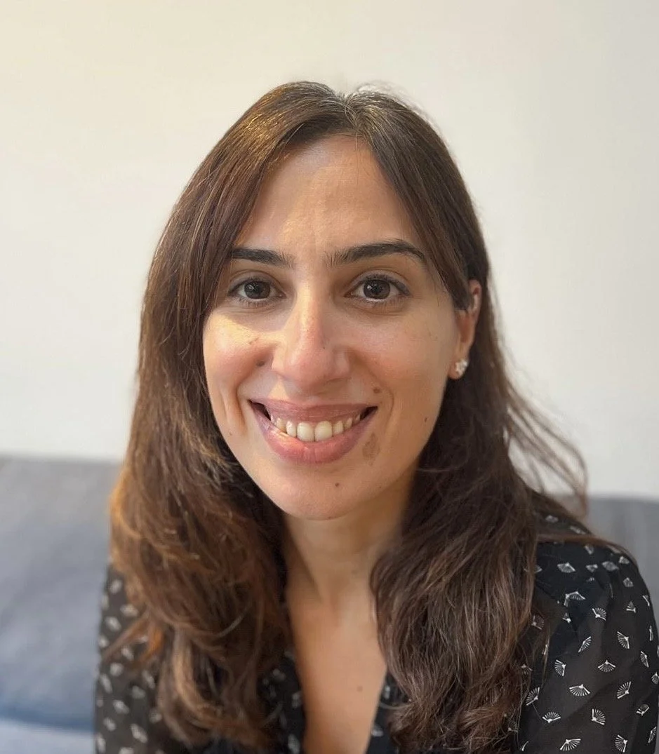 A woman with long brown hair, smiling, wearing a black shirt with white patterns, in front of a neutral background.
