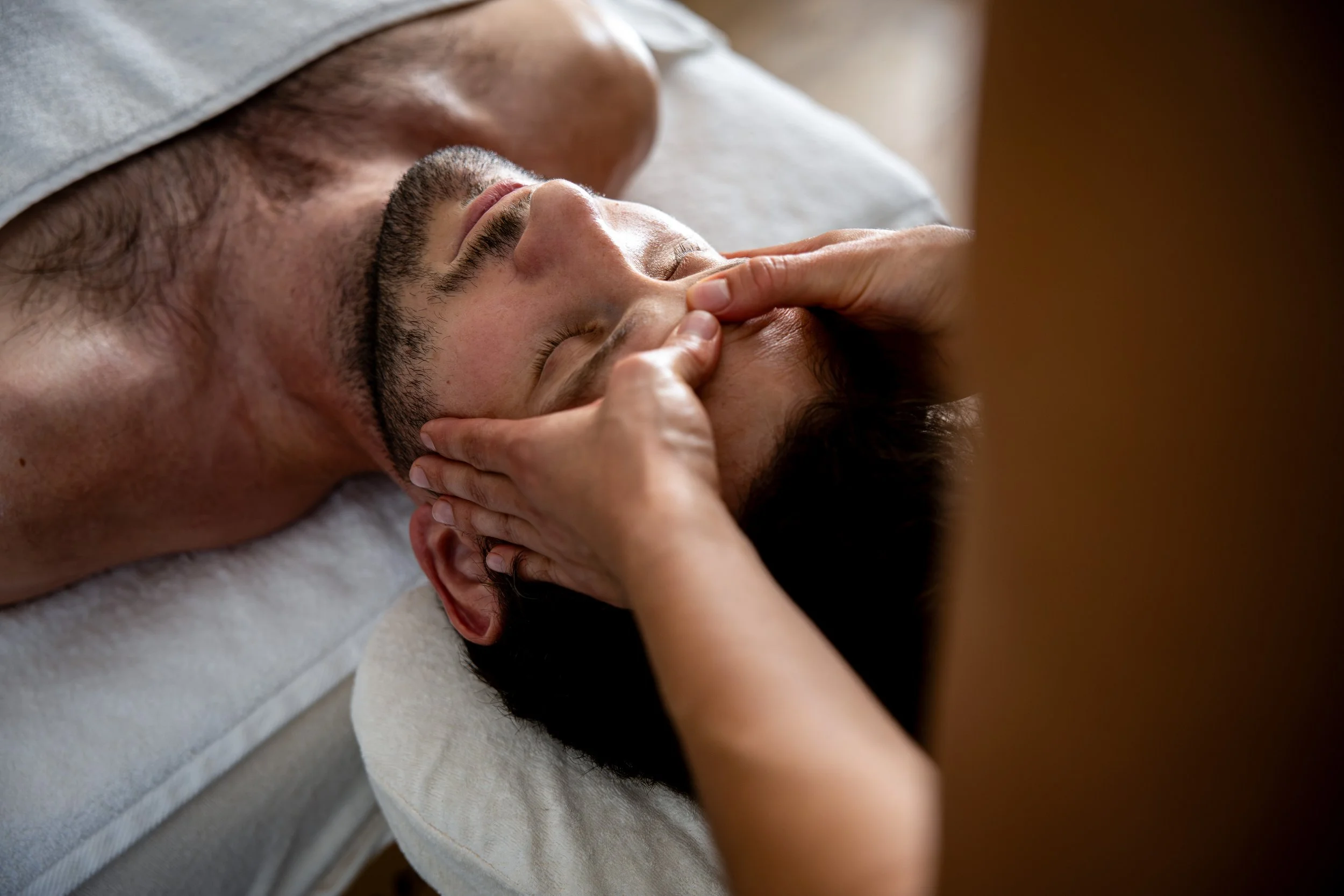 Un homme reçoit un massage du visage, en position allongée. La masseuse effectue des lissages avec ses pouces au niveau de la ride du lion, située entre les sourcils. 