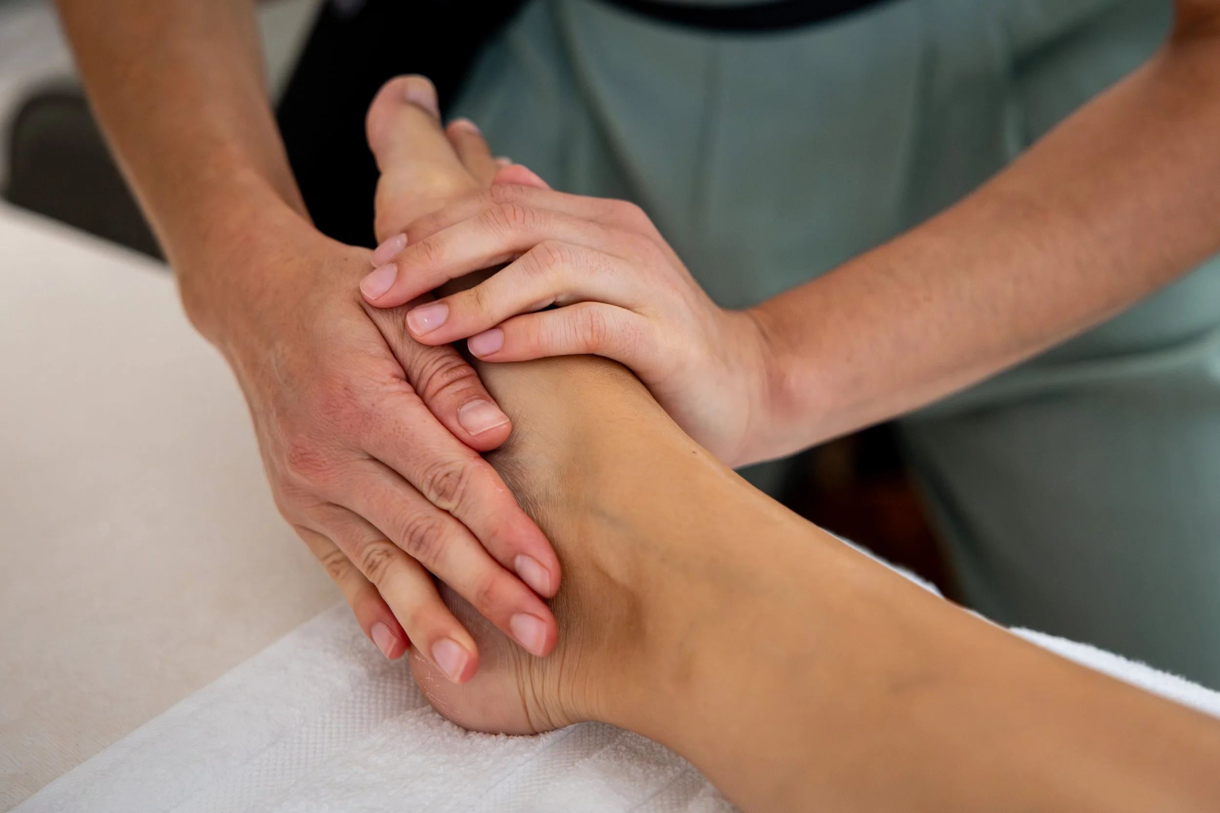 Une personne reçoit un massage du pied par une masseuse, une des mains du thérapeute est sur le dessus du pied l'autre sur le bord interne du pied.