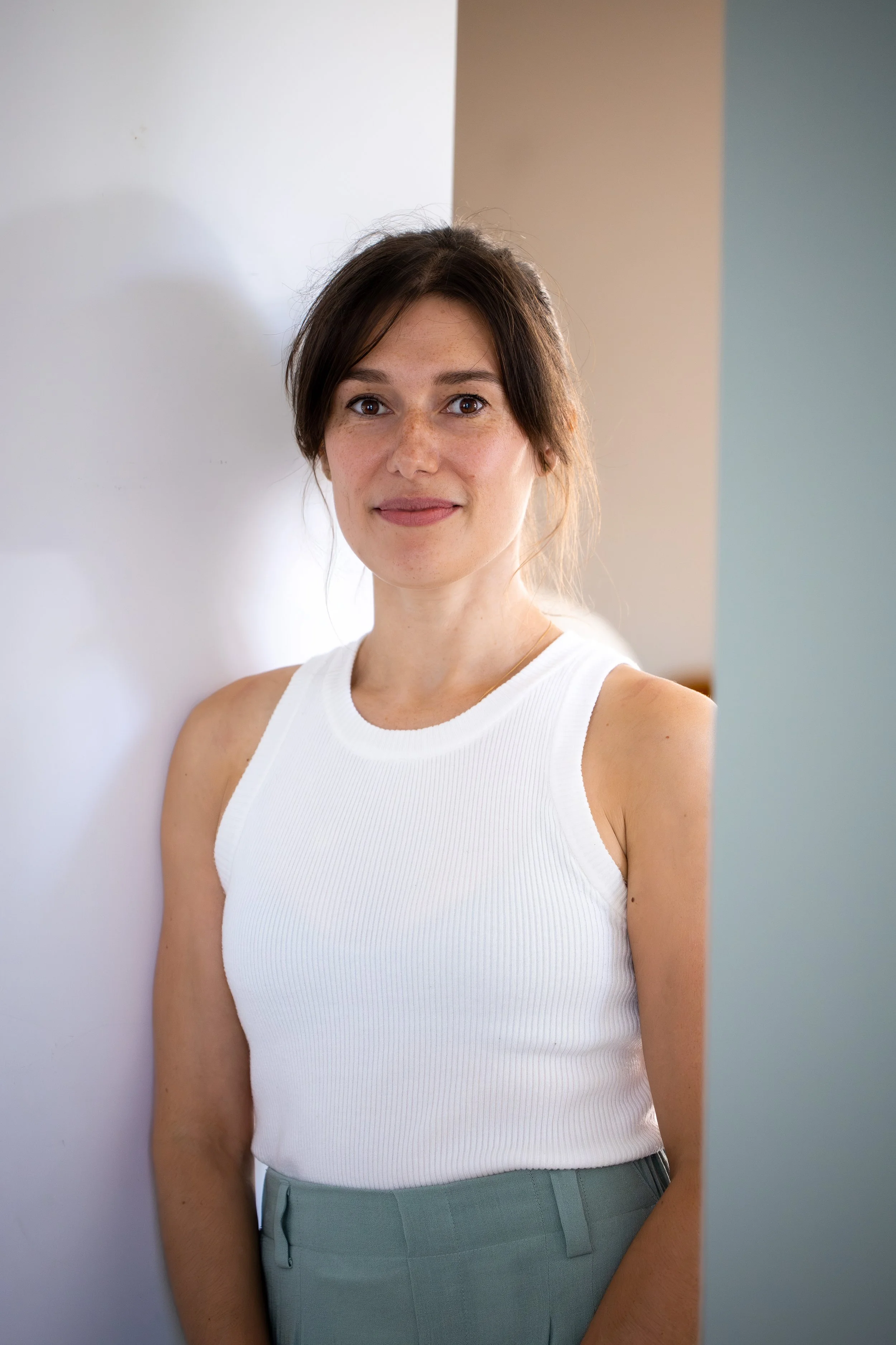 Femme souriante portant un débardeur blanc et un pantalon vert clair, debout entre un mur blanc et un mur beige, regardant vers la caméra.