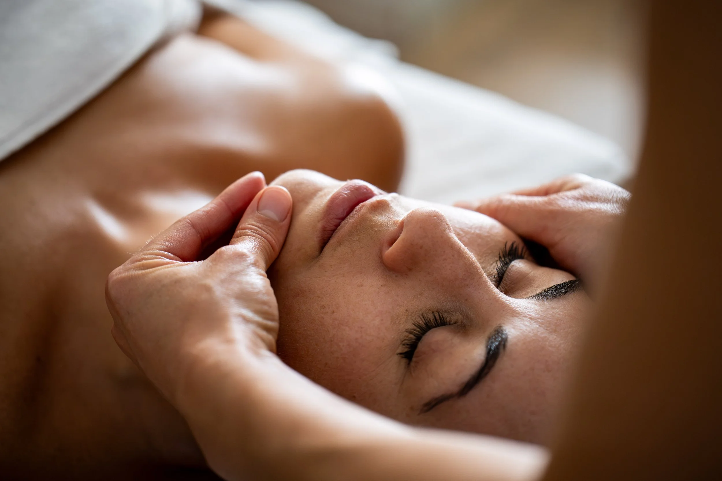Une femme allongée avec les yeux fermés, reçoit un massage du visage, lissages de l'ovale du visage, dans un environnement relaxant.