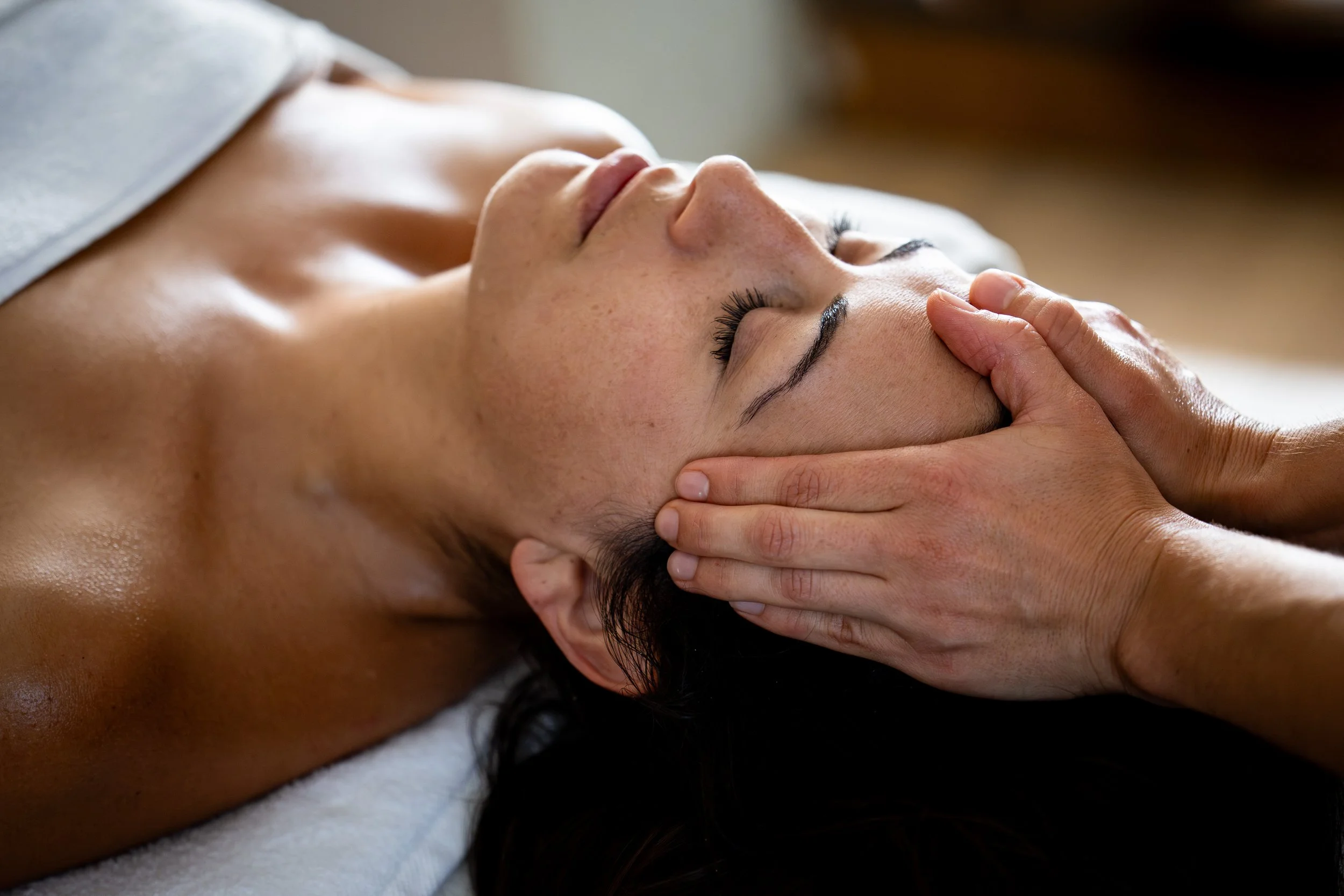 Une personne reçoit un massage du visage avec les yeux fermés, allongée sur une table de massage. Les mains de la masseuse sont posées en casque, à la racine des cheveux de la personne massée. 